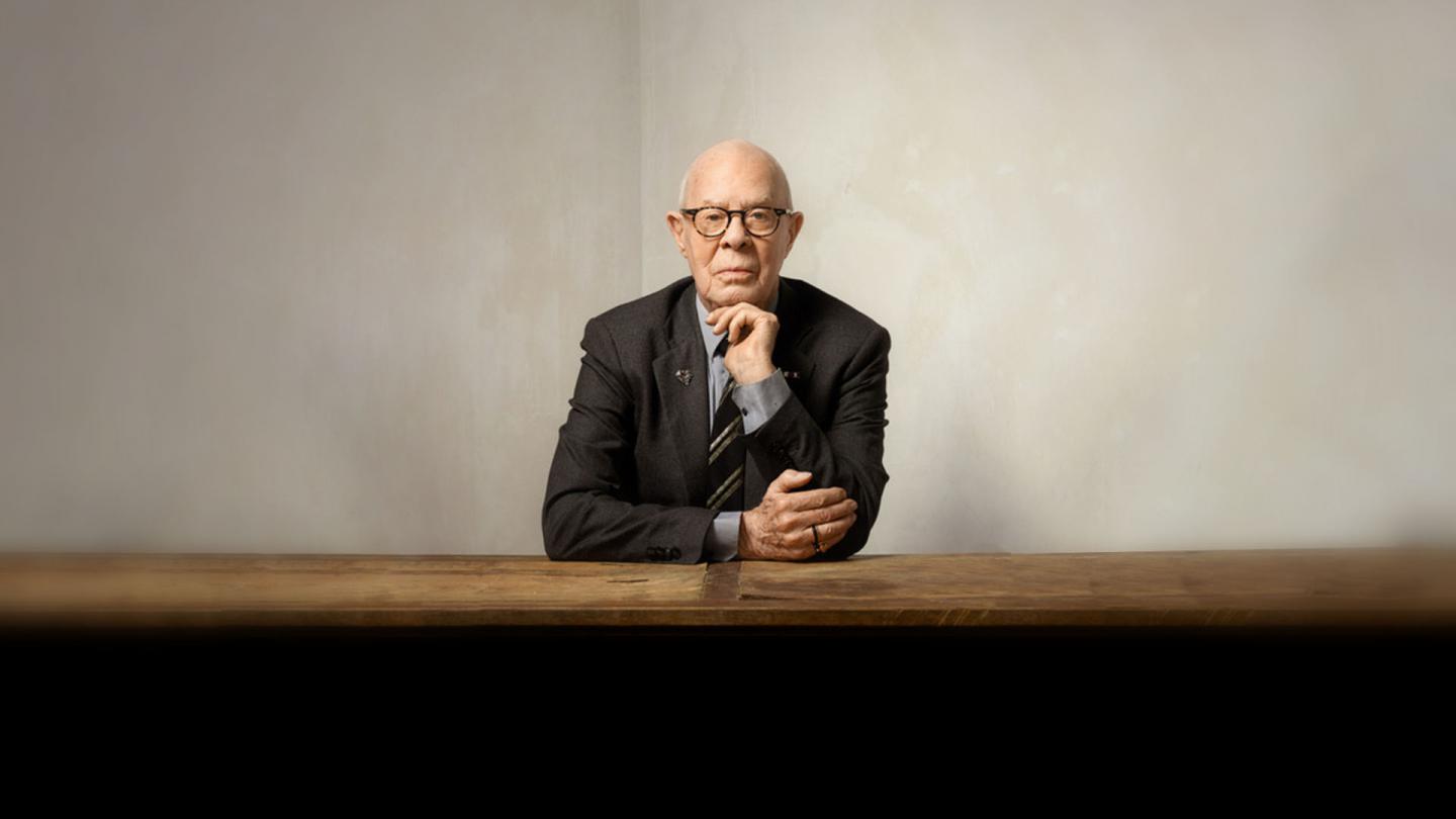 Hans van Manen sat at a wooden table with his chin on one hand wearing a dark suit, grey shirt and black and grey striped tie