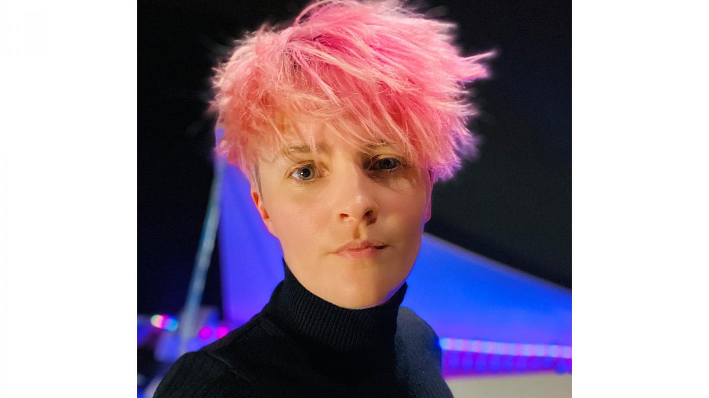 Person with pink hair swept forward, blue eyes, and wearing a black turtleneck top