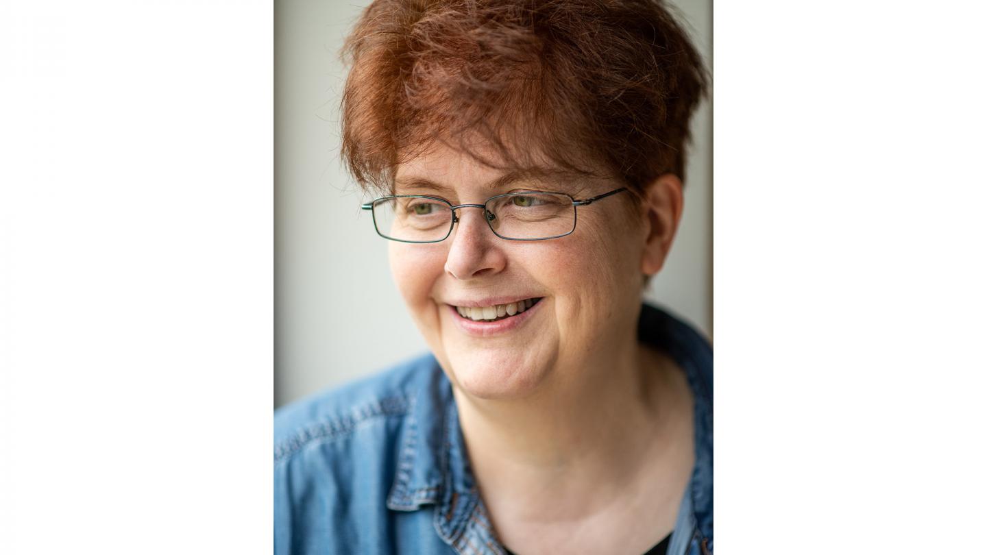 Person with dark red hair, wearing glasses, a denim shirt, smiling.