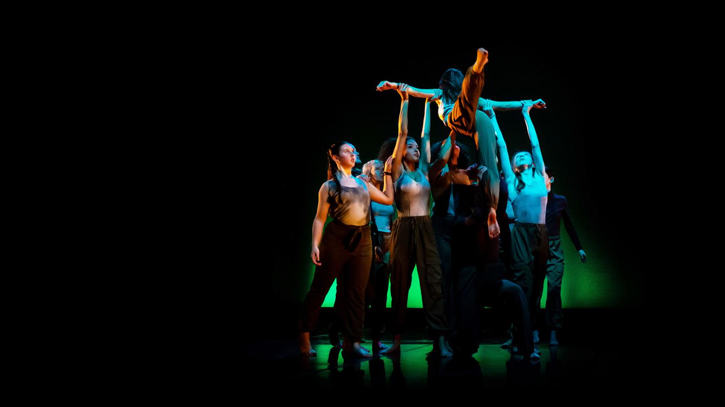 A dancer with leg outstretched in a low atmospheric light being lifted by a group of dancers underneath. 