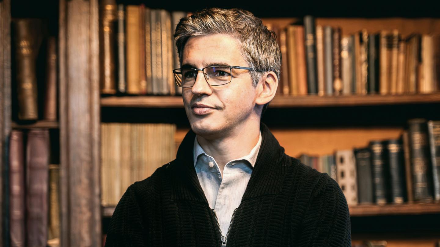 A man stood in front of a wooden bookcase wearing a blue shirt and black jumper. He is wearing glasses and staring off into the distance