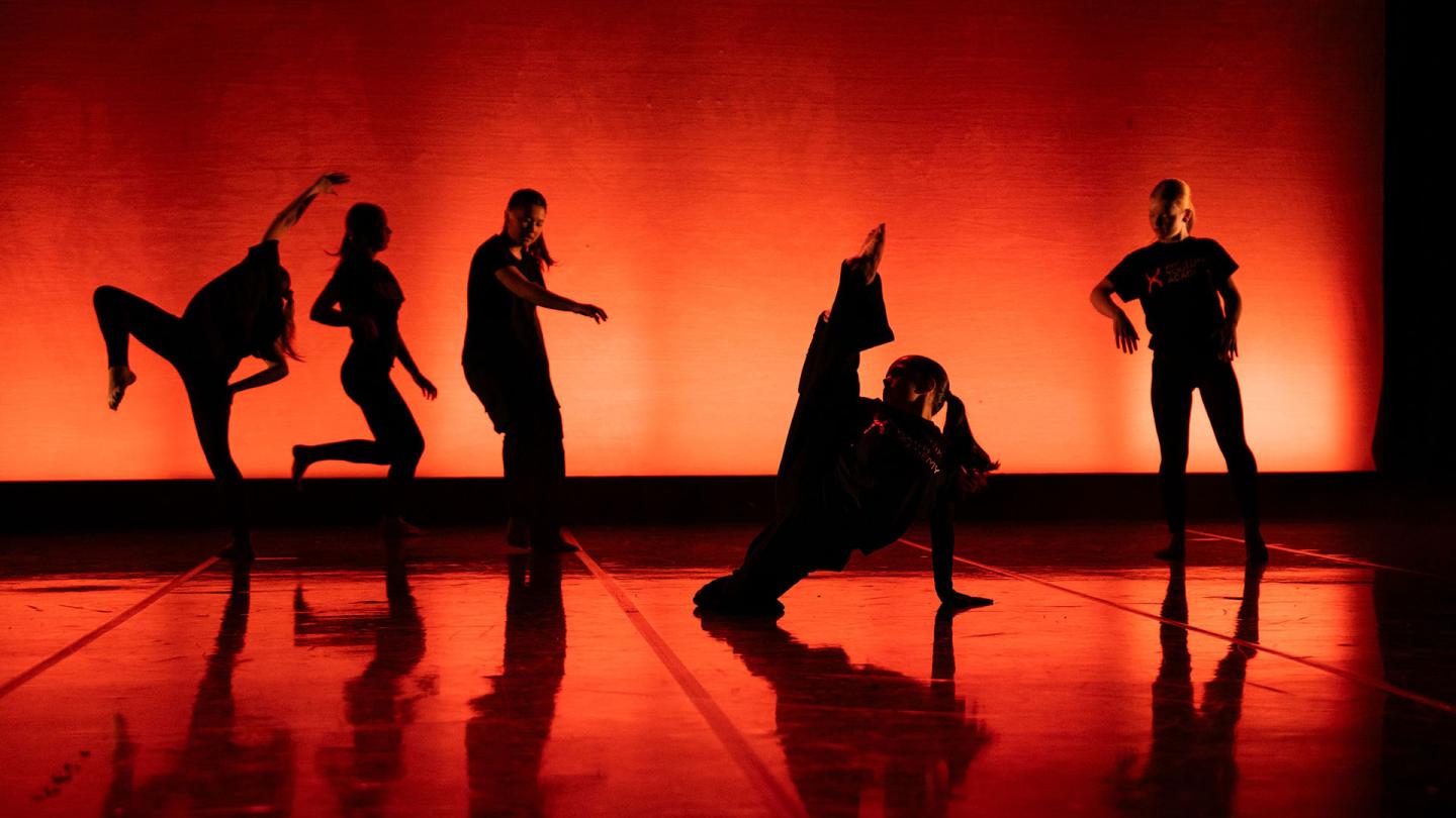 The back is illuminated in red and orange, dancers are closer to us in near silhouette, each in a different but striking pose