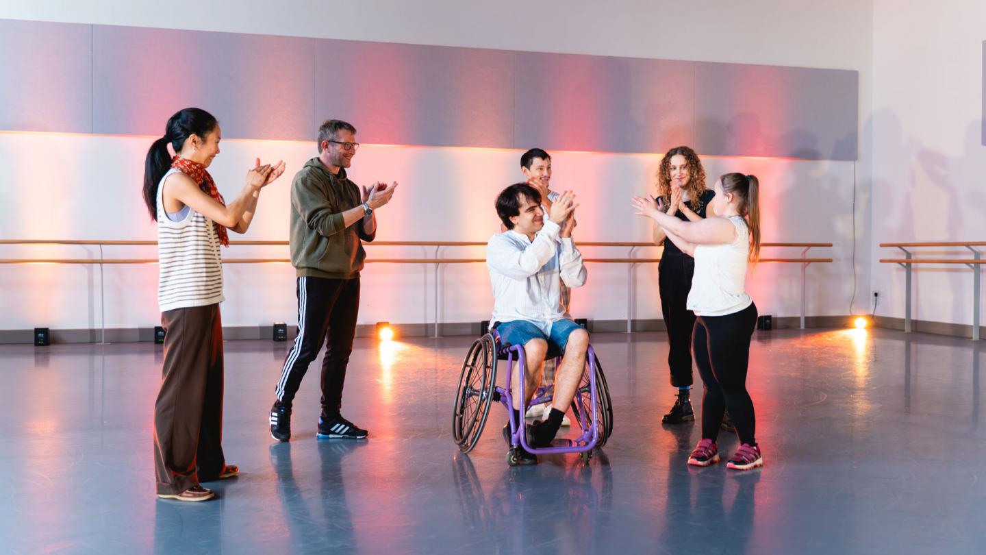 Six people stand infront of warm pink light facing each other, they are applauding.