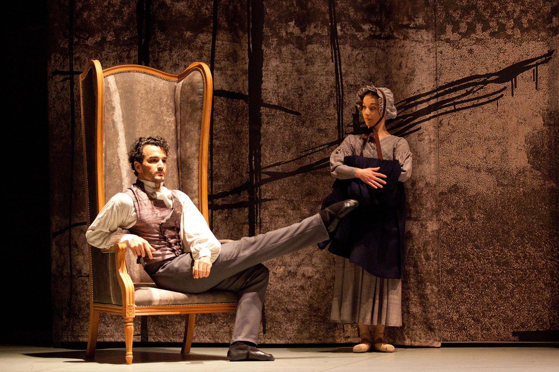 Heathcliff extends his leg while sat in his chair while Mrs Fairfax patiently looks on