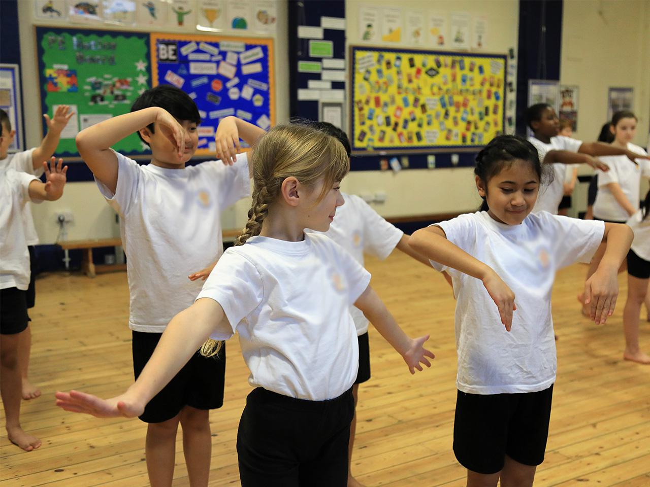 A group of pupils dancing in a school hall. They are holding their arms in different positions: some have their arms outstretched, others and folding them in above or below their shoulders.