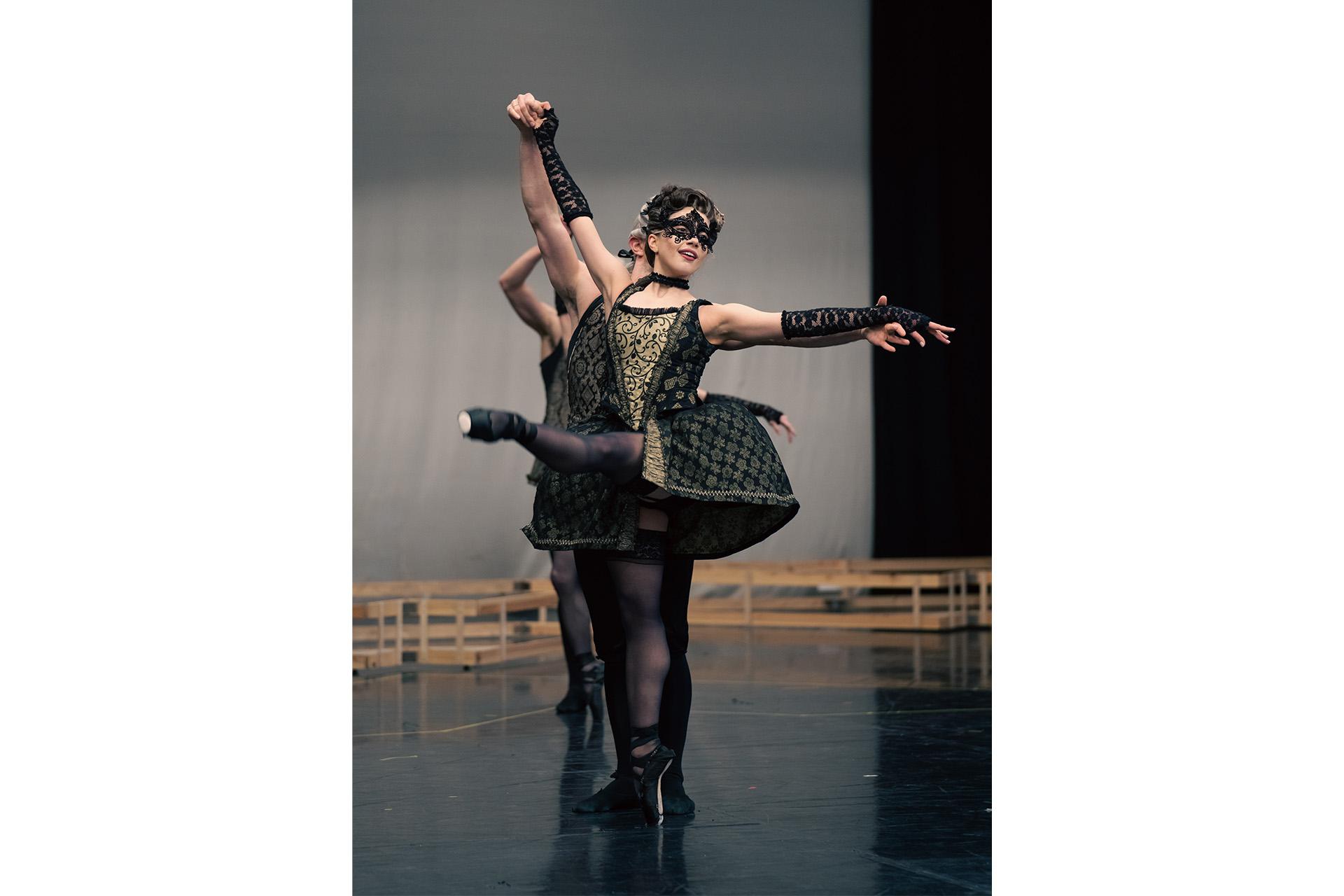 Dancer wearing a black lace mask has foot extended out in front of her.