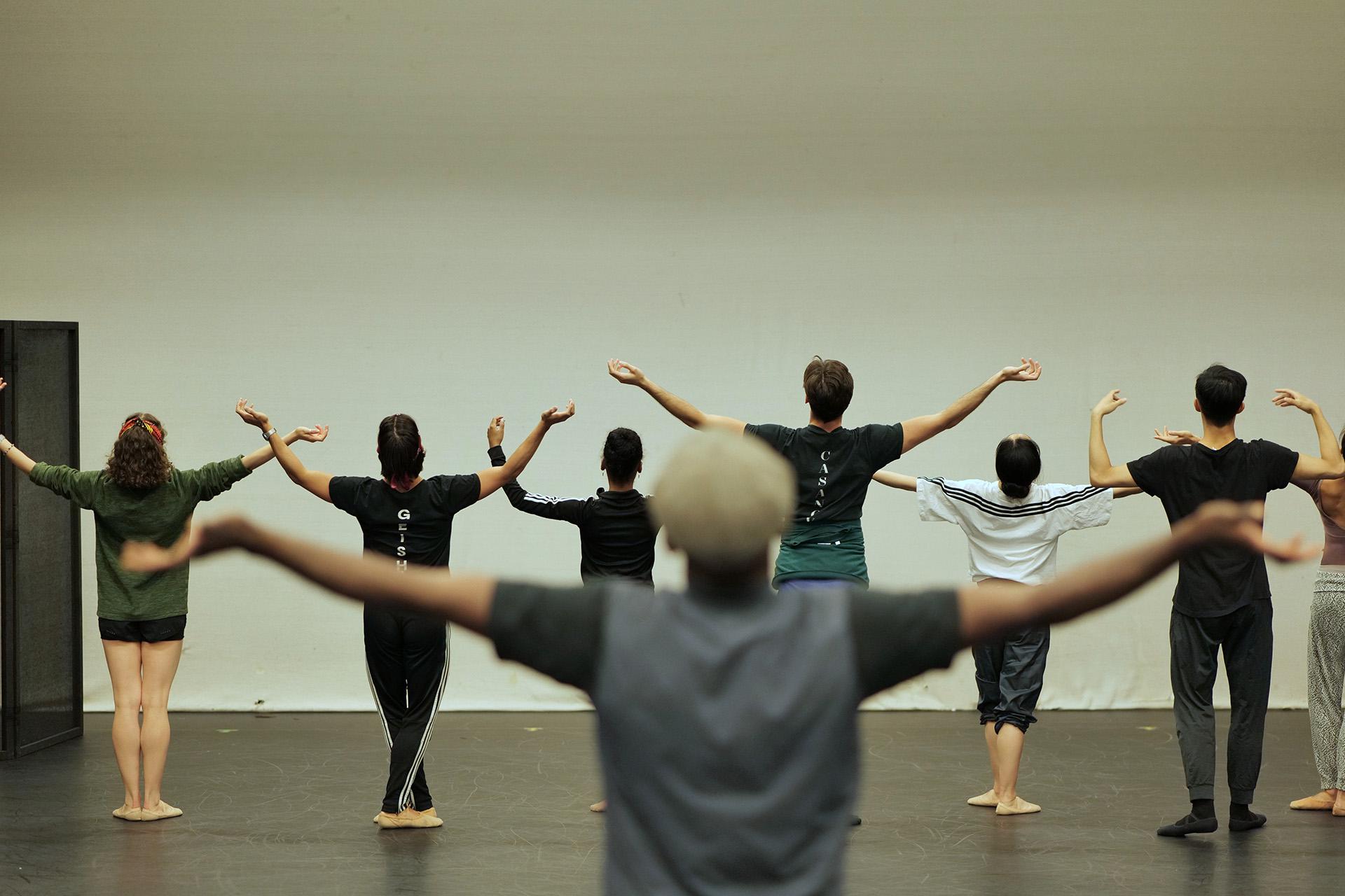 Dancers face a white wall and hold their arms out questioningly