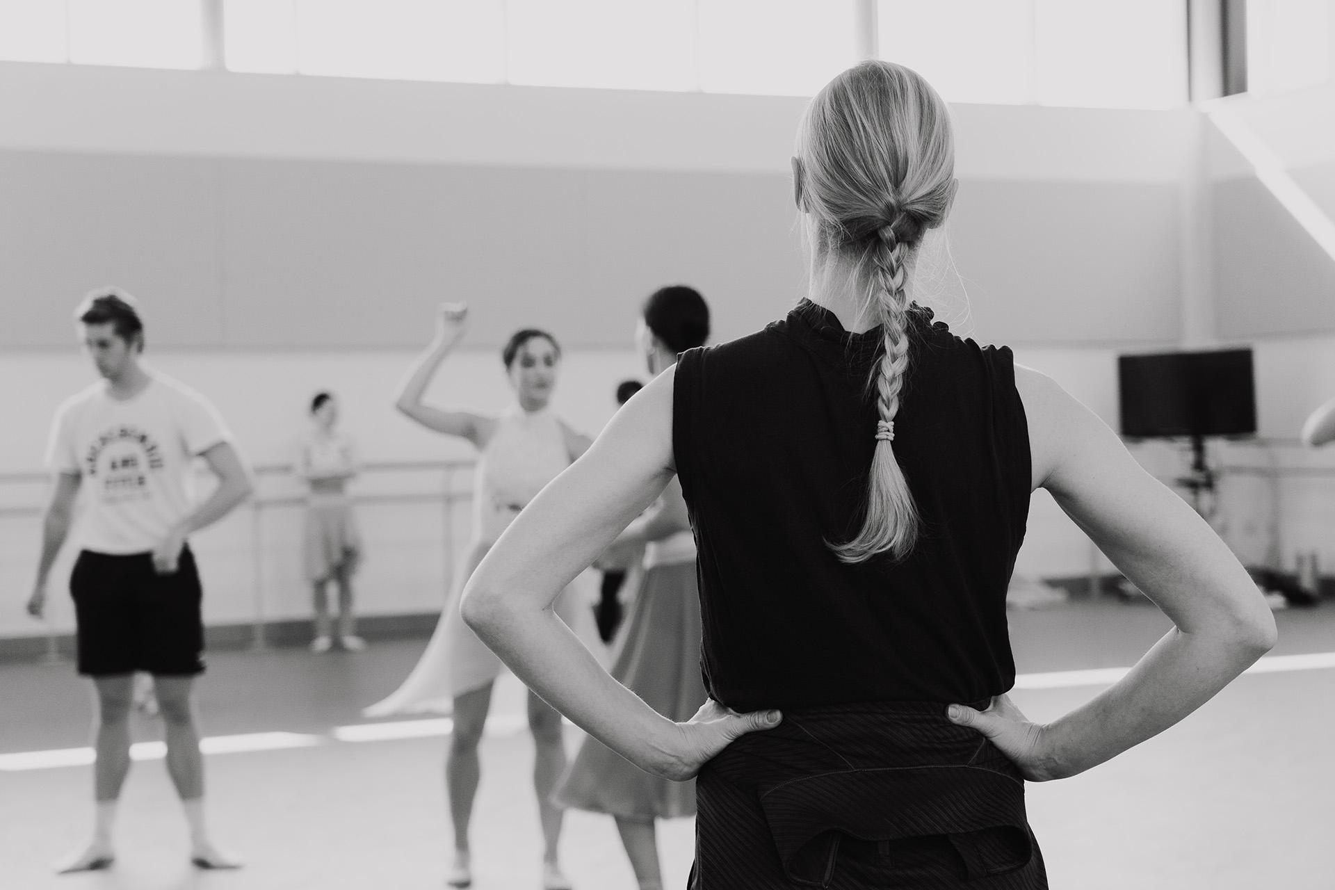 Monochrome image of Stina Quagebeur facing away from the camera with her hand on her hips watching dancers rehearse