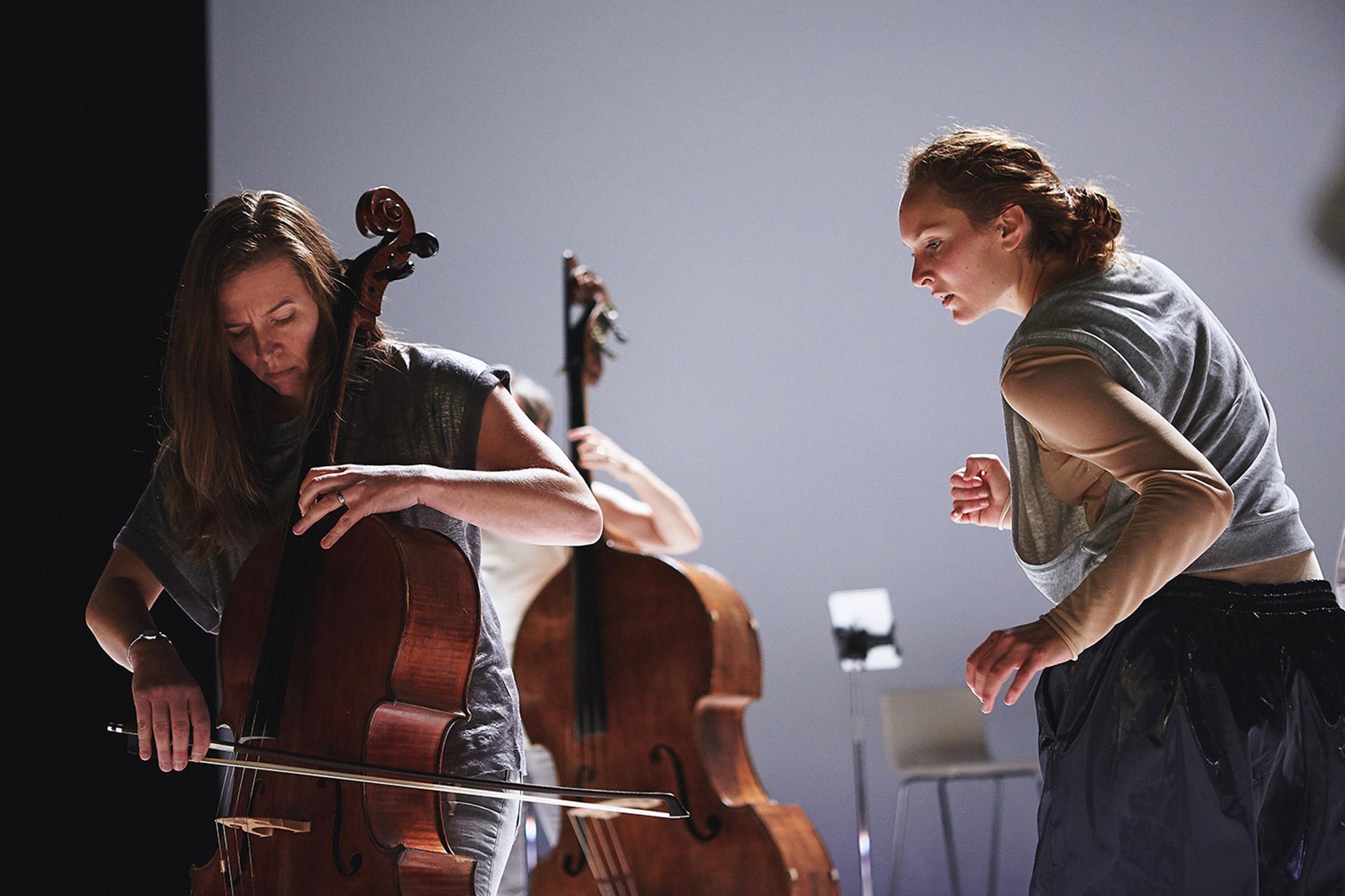 A cellist intently plays her instrument while a person in blue trousers, grey top, and hair tied back looks on