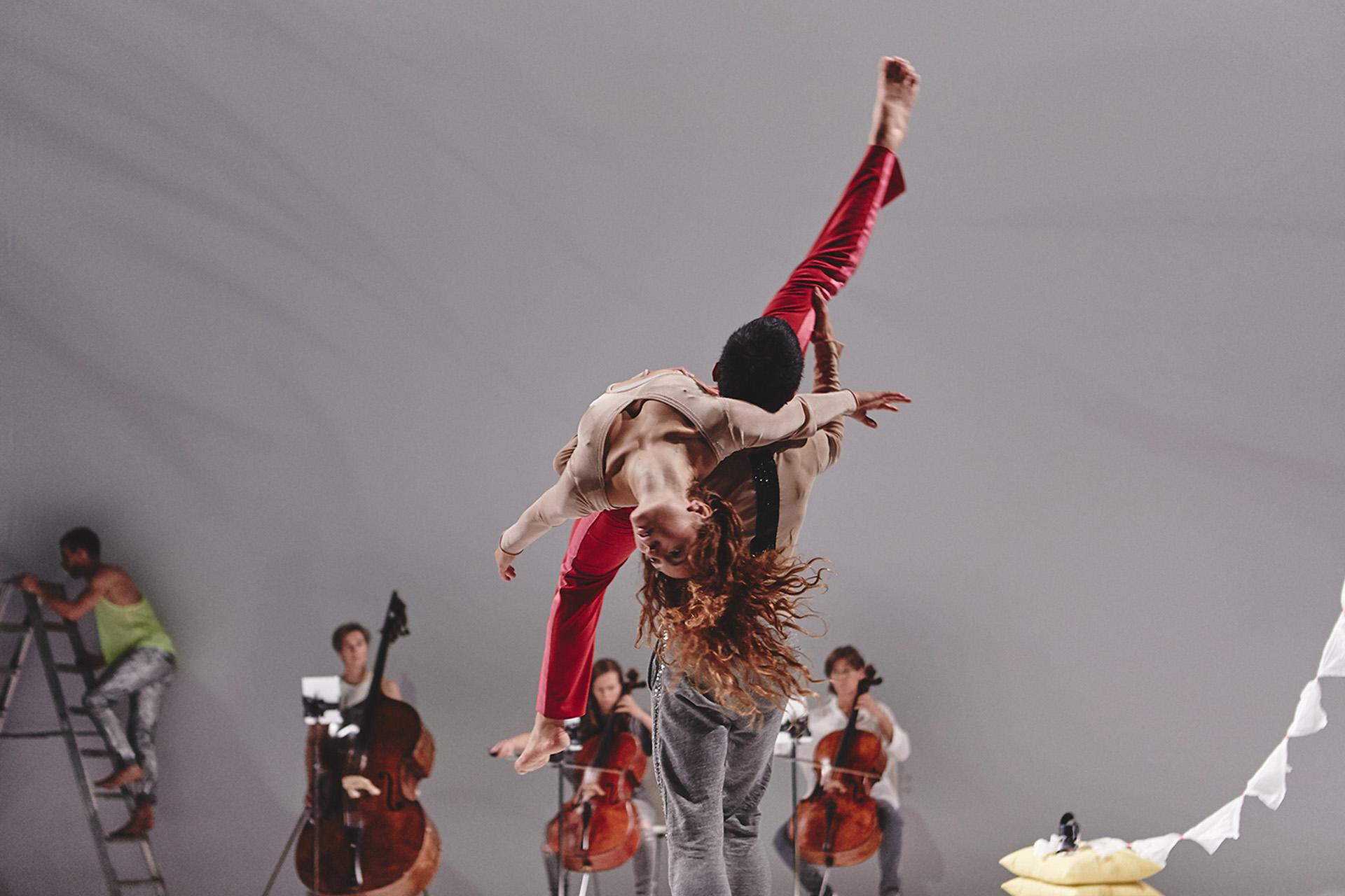a dancer facing away from the camera holds a person wearing vivid red trousers on his shoulders, in the background a man climbs a stepladder and three people are sat playing their large stringed instruments
