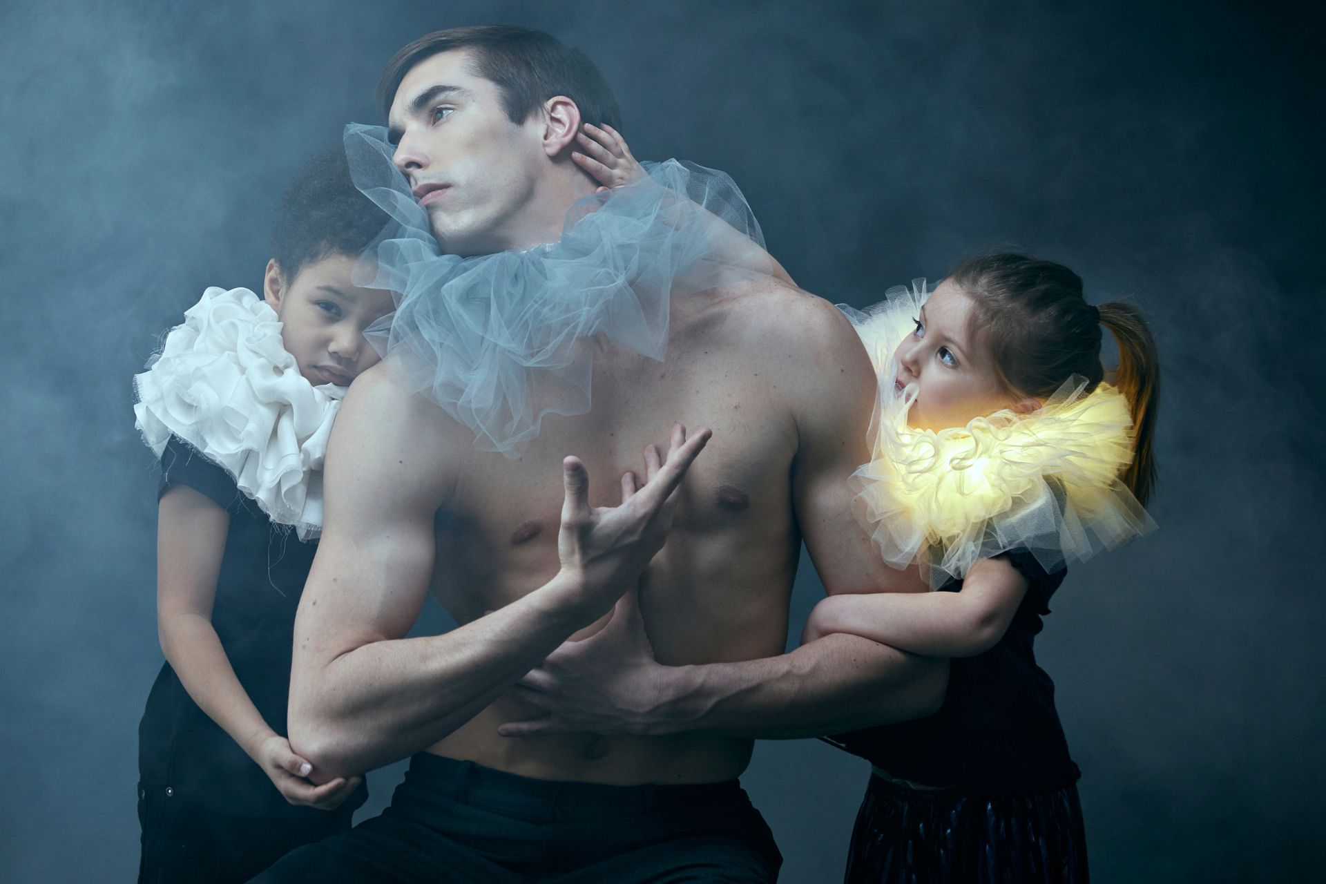 A man and two young children wearing colourful toule around their necks. They are surrounded by smoke with a dark grey background behind them.