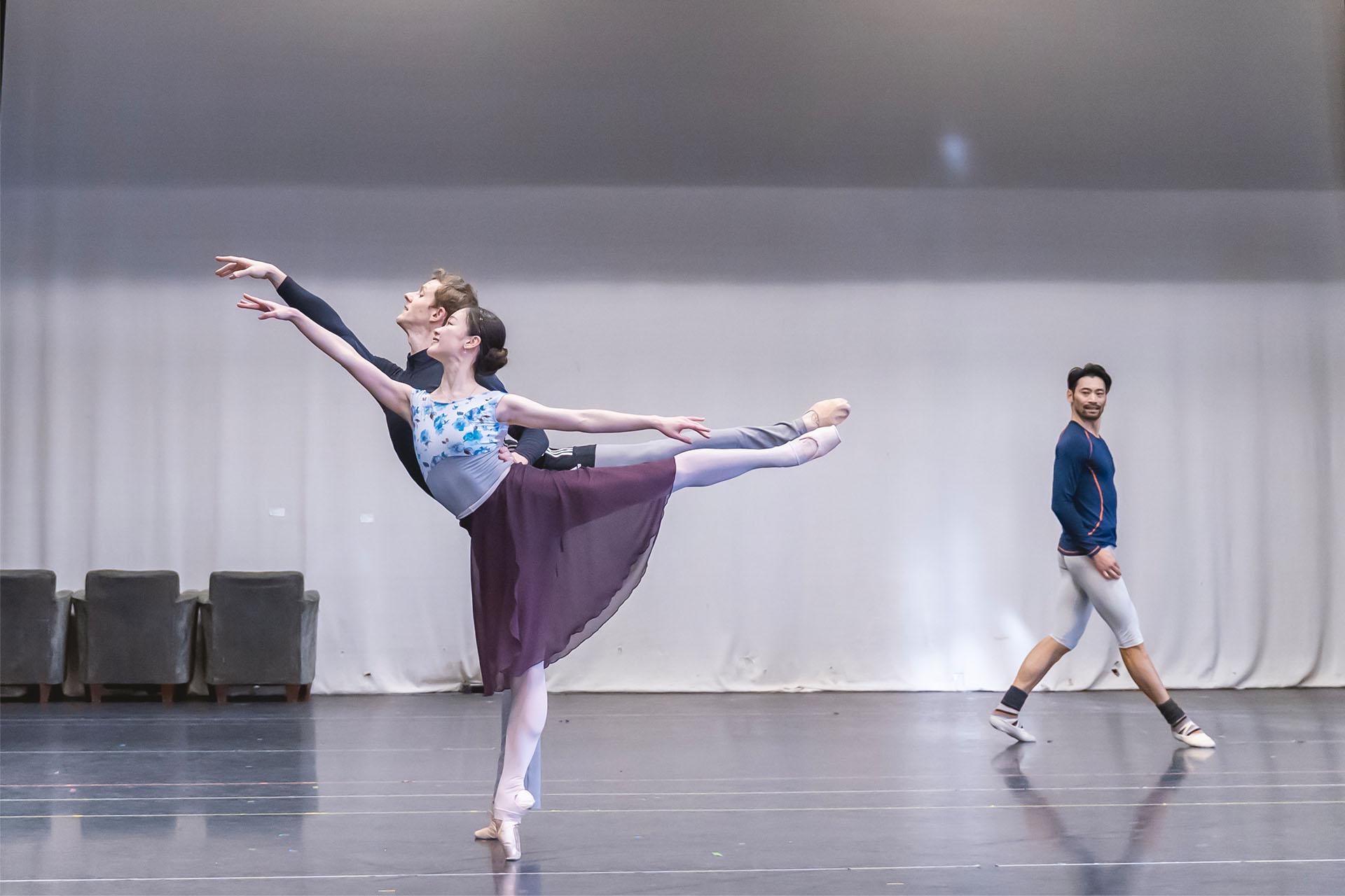 Two dancers stand arabesque, the woman en pointe, while another dancer strides in the background watching them