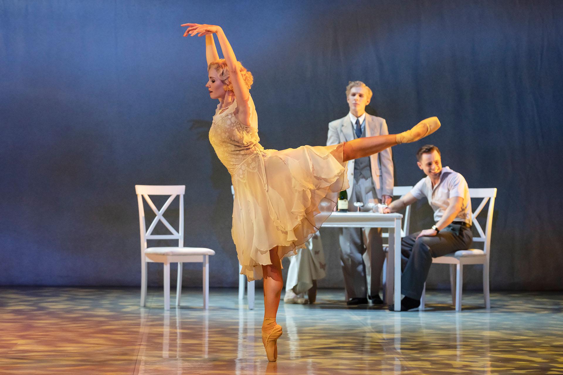 Blonde female dancer in arabesque wearing a flowing white dress while a friend looks on