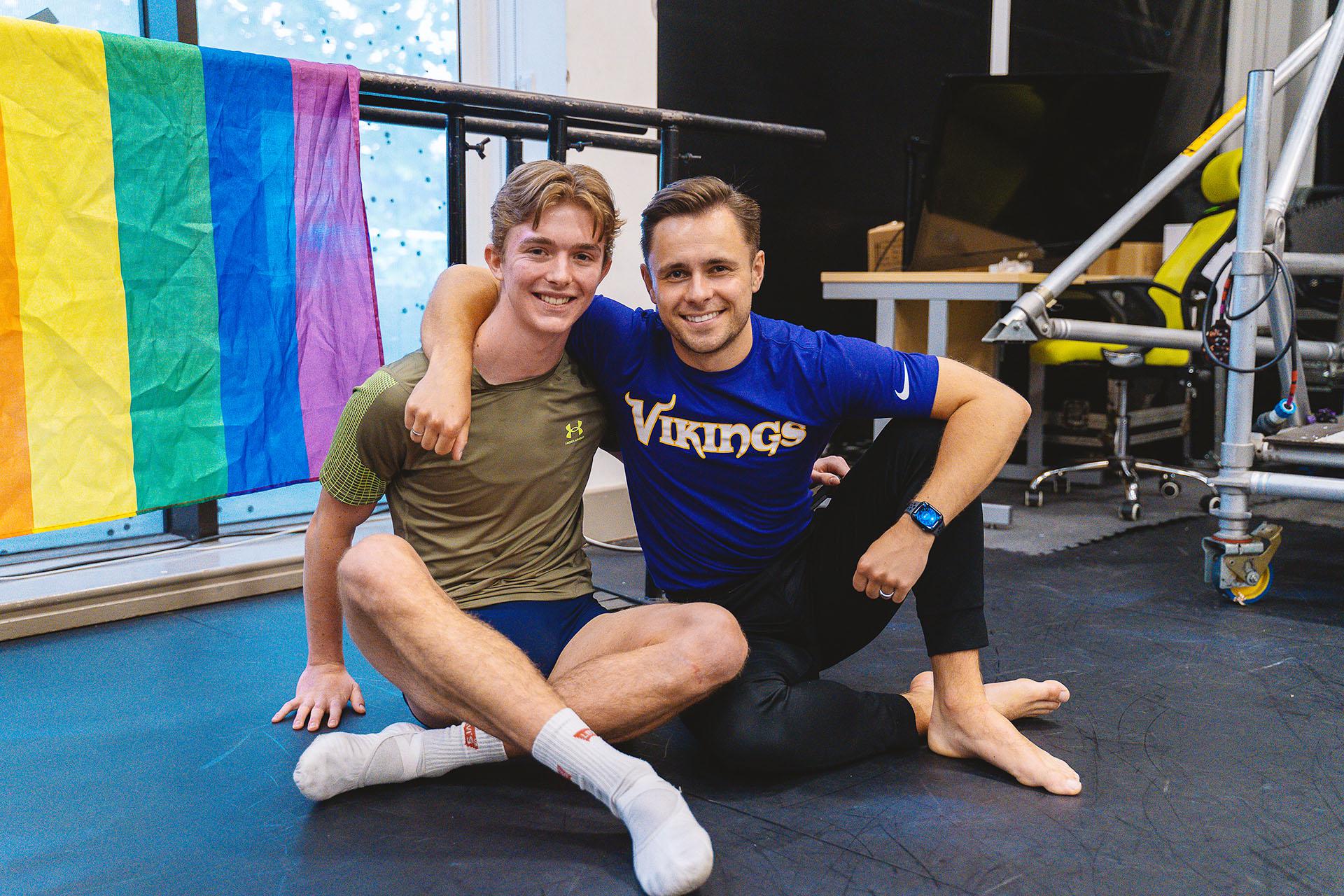 Two dancers sat on the floor, a Pride flag behind them, smiling directly at the camera