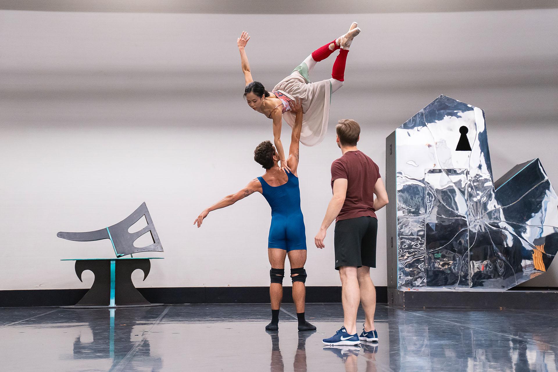 In rehearsals for Beauty & the Beast and male dancer holds a female dancer by her waist above his head with one arm. She has one hand on his shoulders and the other arm, and legs, are raised.