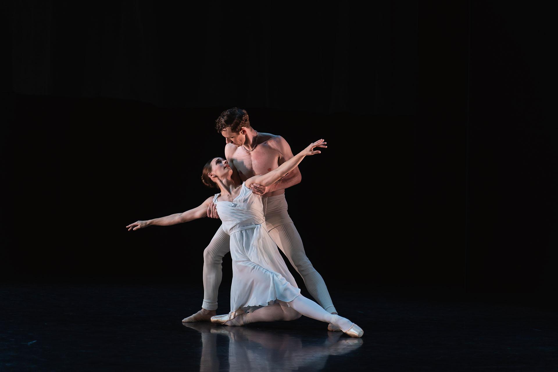 Dancers in white, one standing and holding the other by their arms as they are close to the ground, on knee bent and the other leg stretched out front