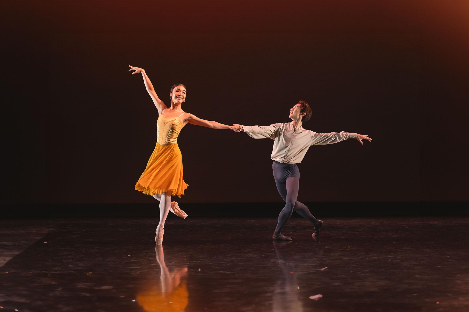 Female dancer in a orange skirt smiling as she stands on one foot en point, one leg bent at the knee behind her and an arm raised above her head while the other is held by a male dancer in white and grey