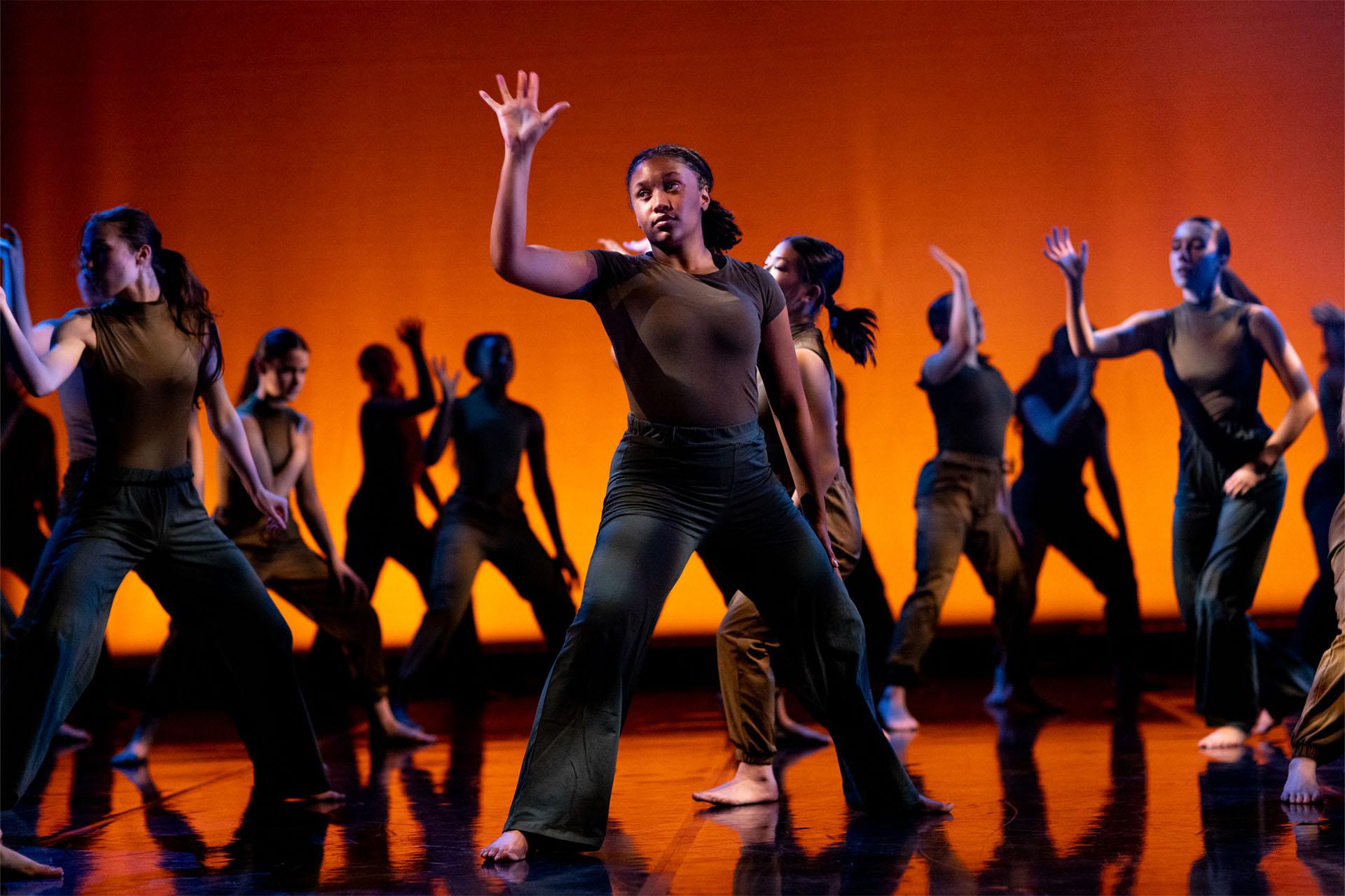 Large group of dancers ion stage, the focus is on a woman standing strong with her hand raised with fingers spread out.
