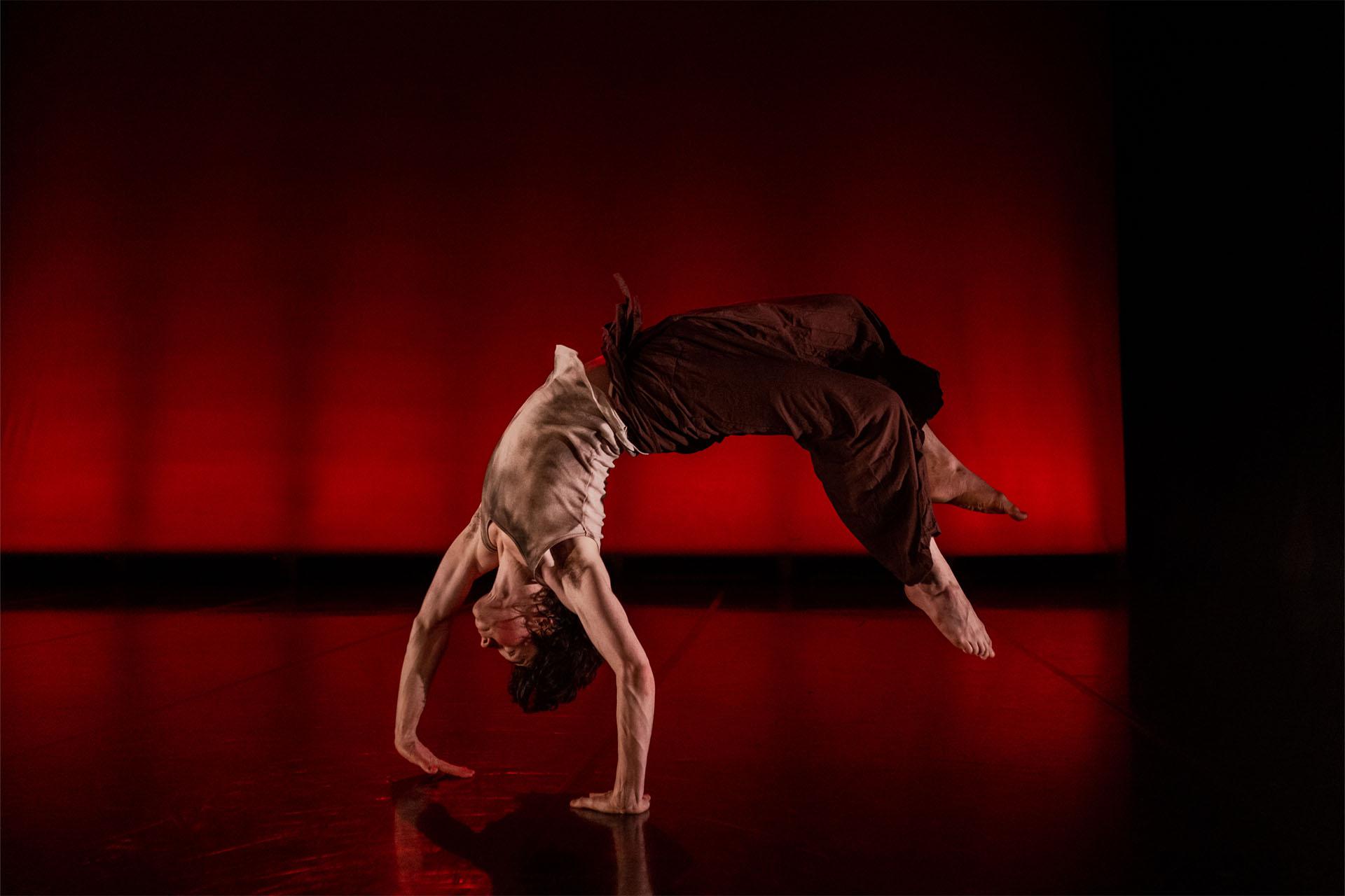 Against a red background, a Senior of the Phoenix Youth Academy mid backflip with his hands on the floor , his back arched and feet just above the ground