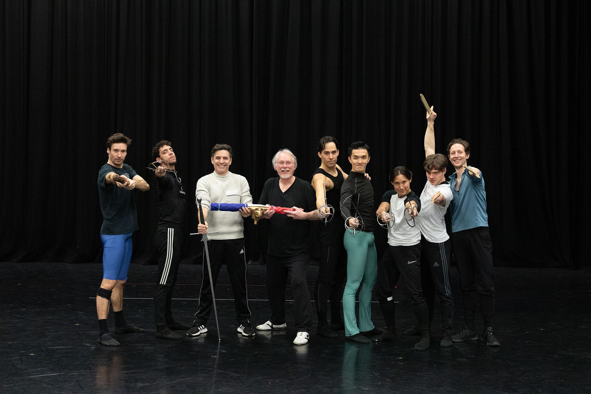 Against a black backdrop the fight director Jonathan Howell, rehearsal director Daniel de Andrade, male dancers line up for their photo after a rehearsal session with staffs and swords