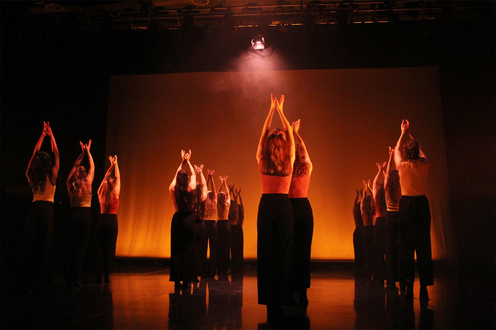 Under orange light, a group of young dancers standing tall and reaching above their head.