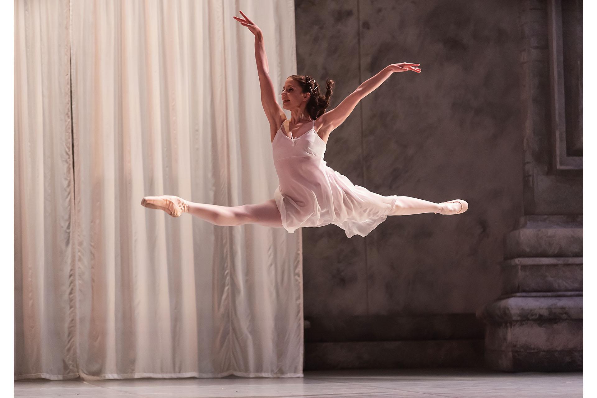 Dancer wearing white flies across the stage, leaping in a grande jete