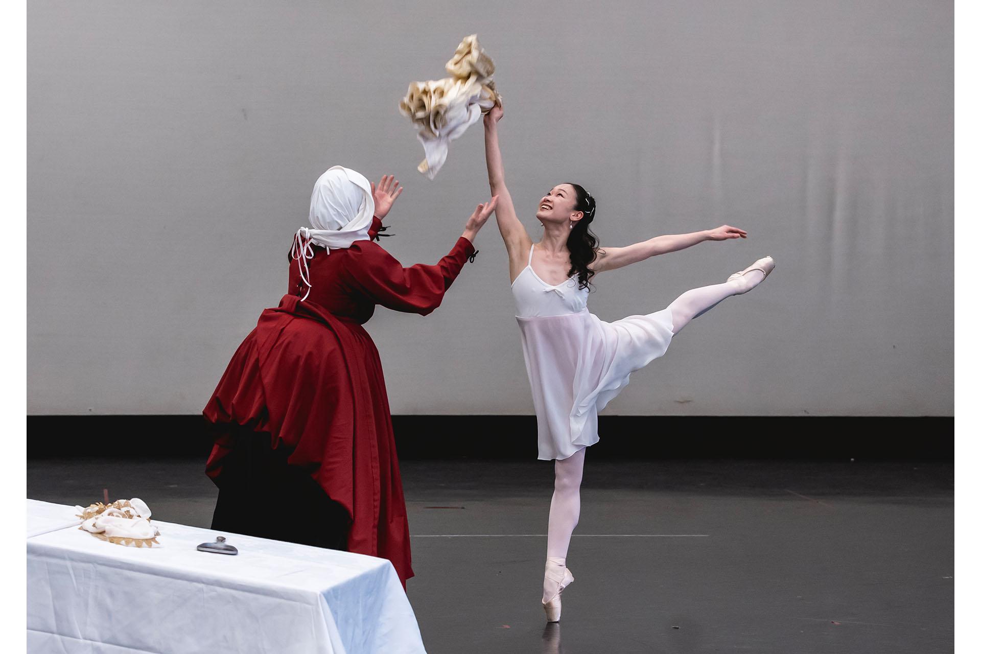 A dancer dressed all in white and grinning stands on one pointed foot and holds a doll above her head while another with her back to us and dresses in red pleads with her to stop playing