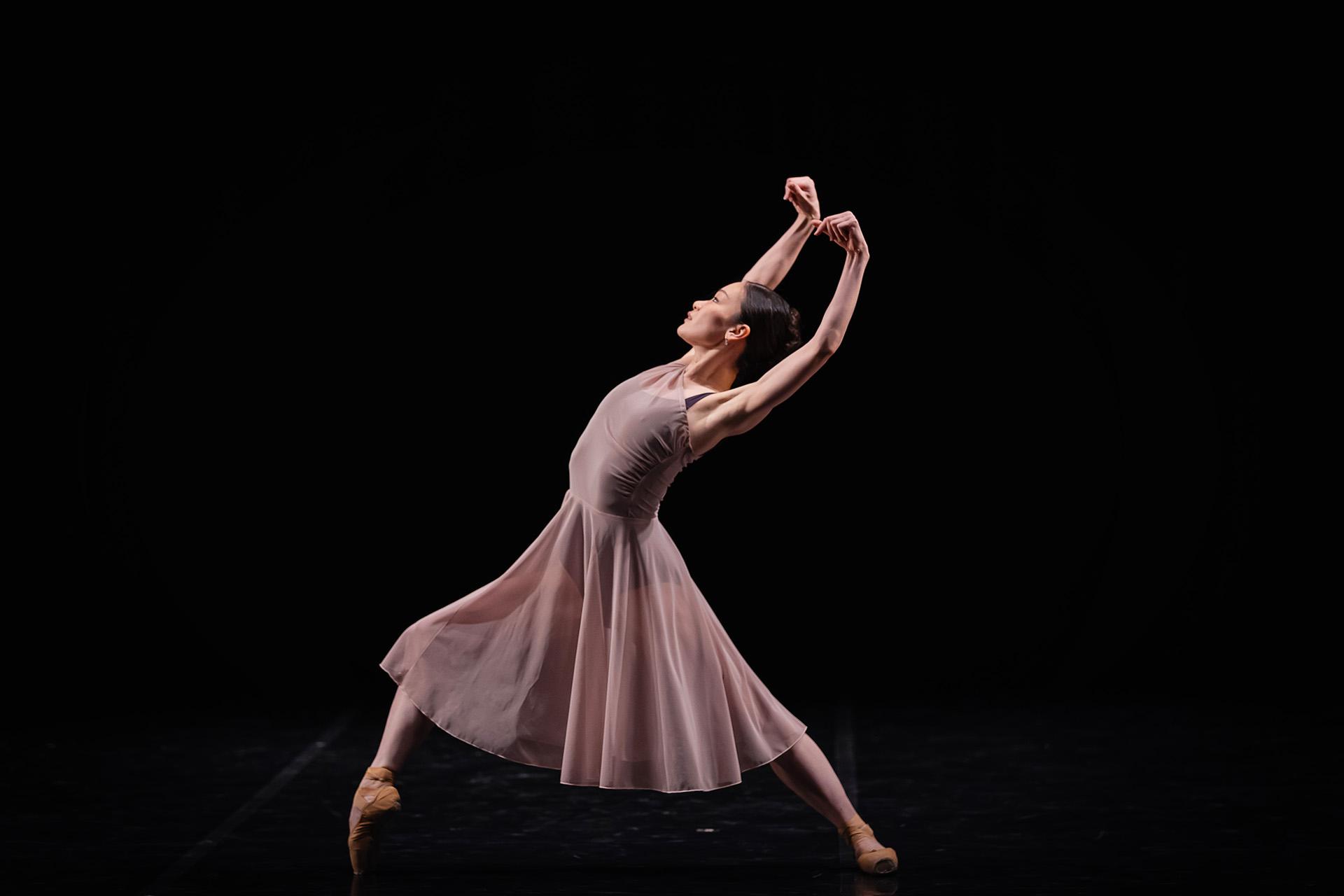 Dancer in a lilac skirt and top stands with arms above her head
