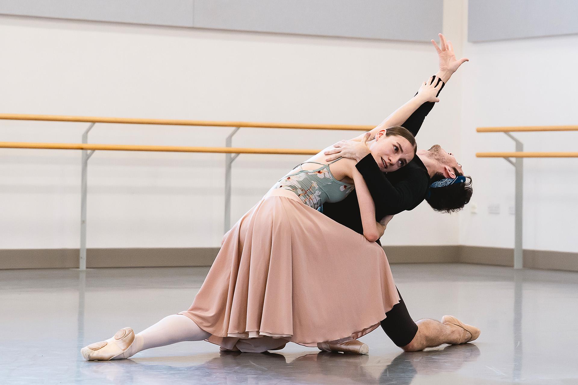 Two dancers in rehearsal on the floor, both kneeling, one resting on the body of the other as he leans back