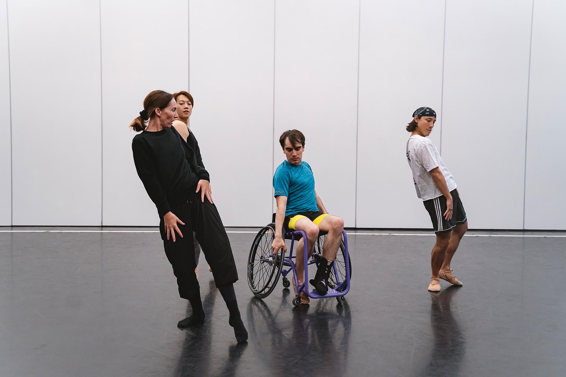 A disabled dancer in a wheelchair poses intently with two dancers and the choreographer each holding a strained pose by him
