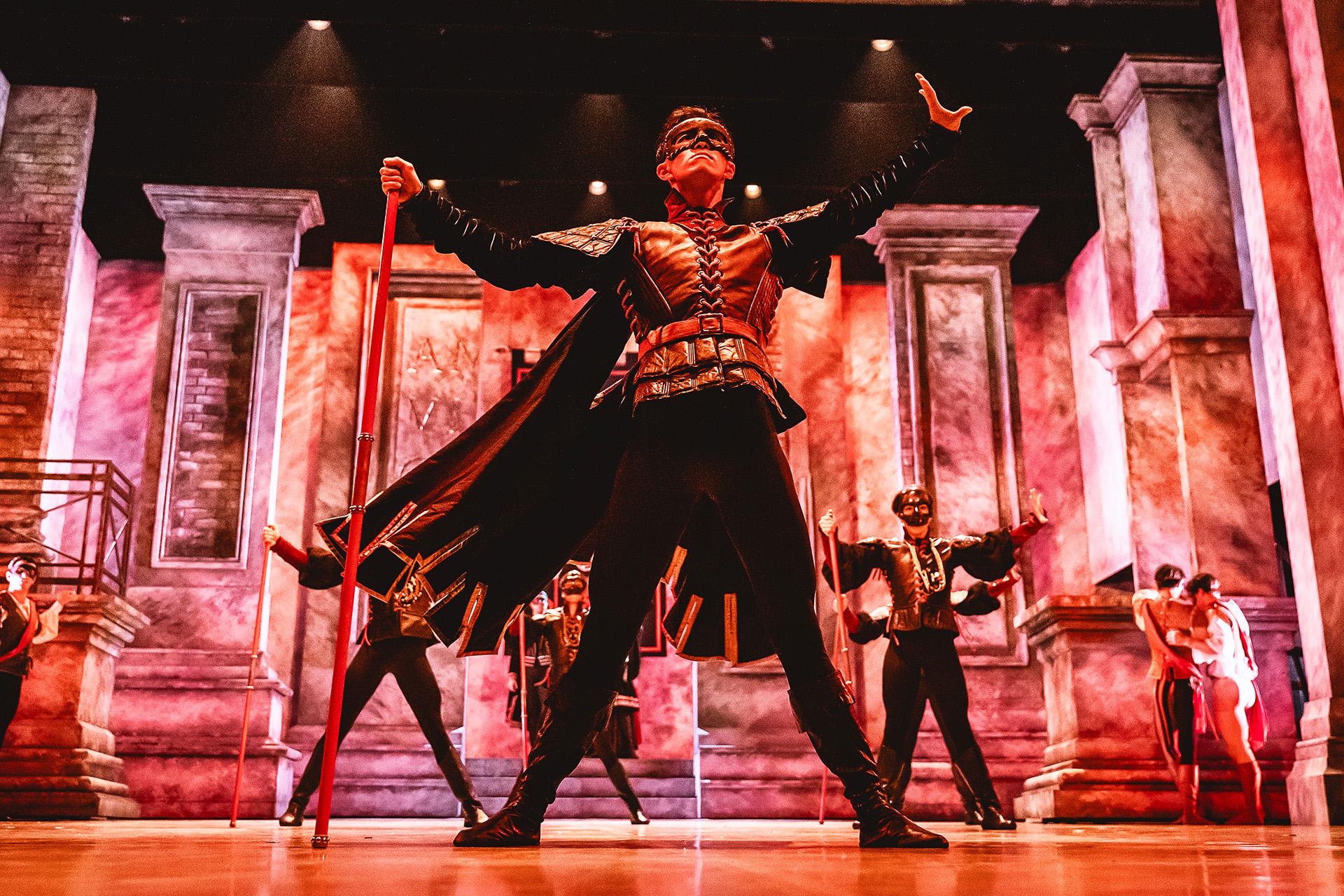 Male dancer wearing a masque on his face, his legs spread firmly apart, holding a red staff in his hand, one end on the floor