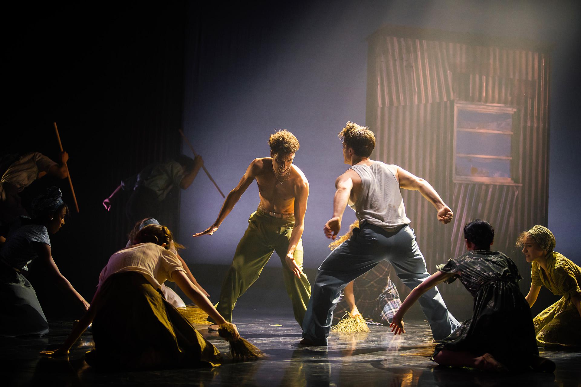 Two dancers, one bear chested in green trousers, the other in blue trousers with a white vest, fighting