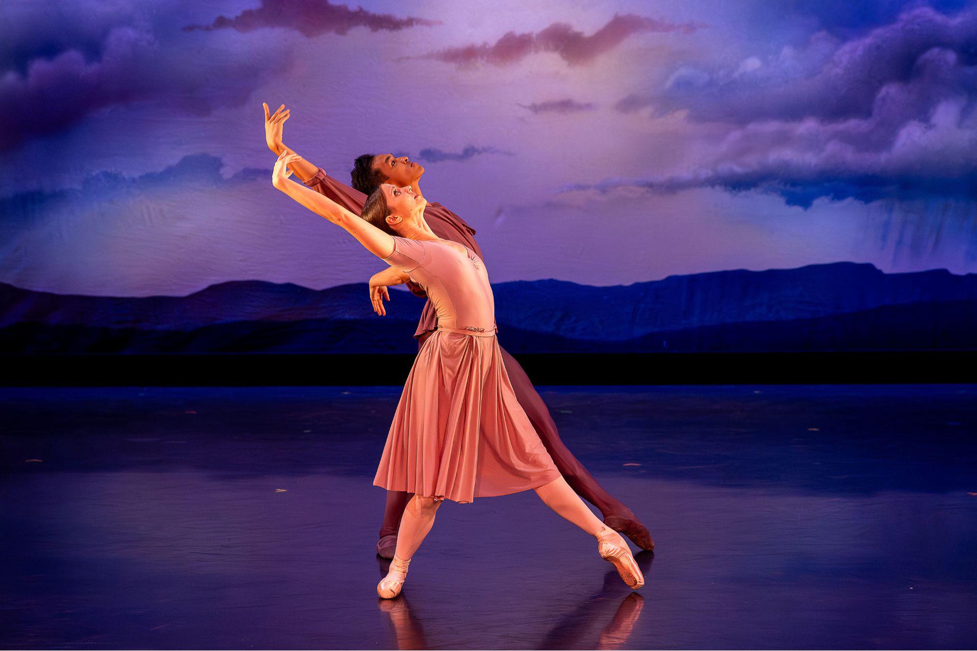 Two dancers against a cloudy background move to the right while leaning backwards and lifting their arm above their heads.