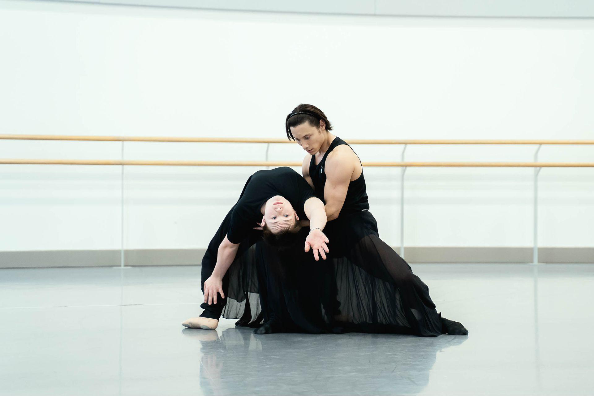 In a rehearsal studio, a dancer kneels as another dancer stretches their back over their bent knee.