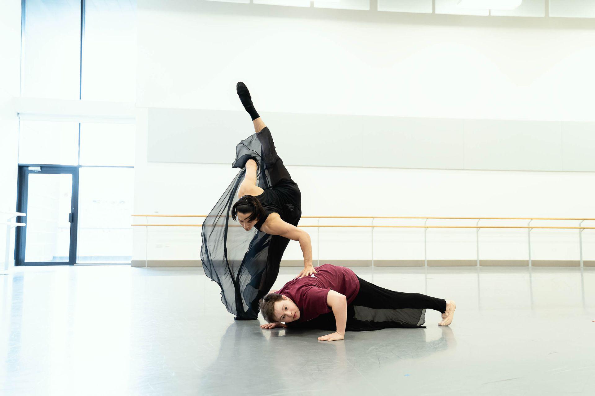 A dancer lies in a press up position as another dancer wearing veiled trousers lifts their leg over them.