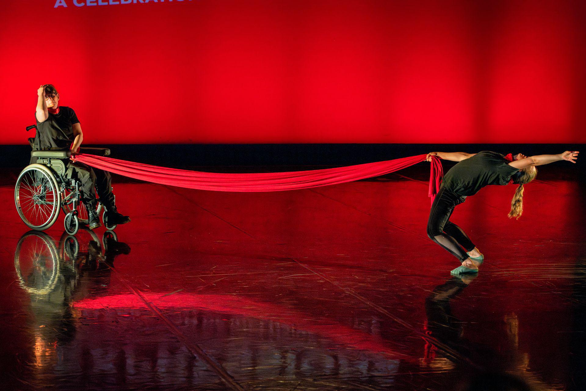 A dancer in a wheelchair holds a long red piece of fabric against a red backdrop. The other end of the fabric is held by another dancer who leans backwards on the other side of the stage.