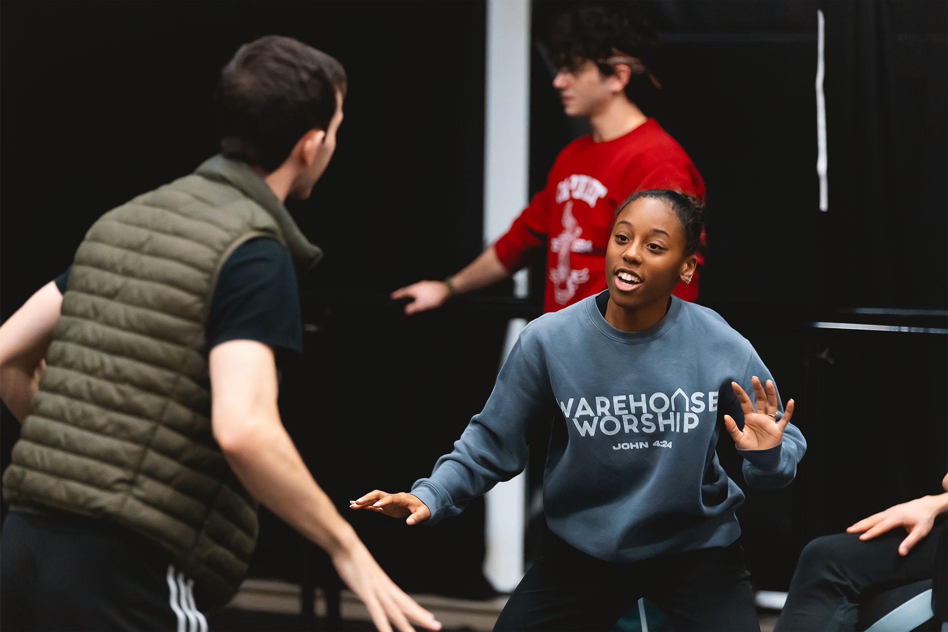 Two dancers in the rehearsal room, the person facing us smiling with her arms waving
