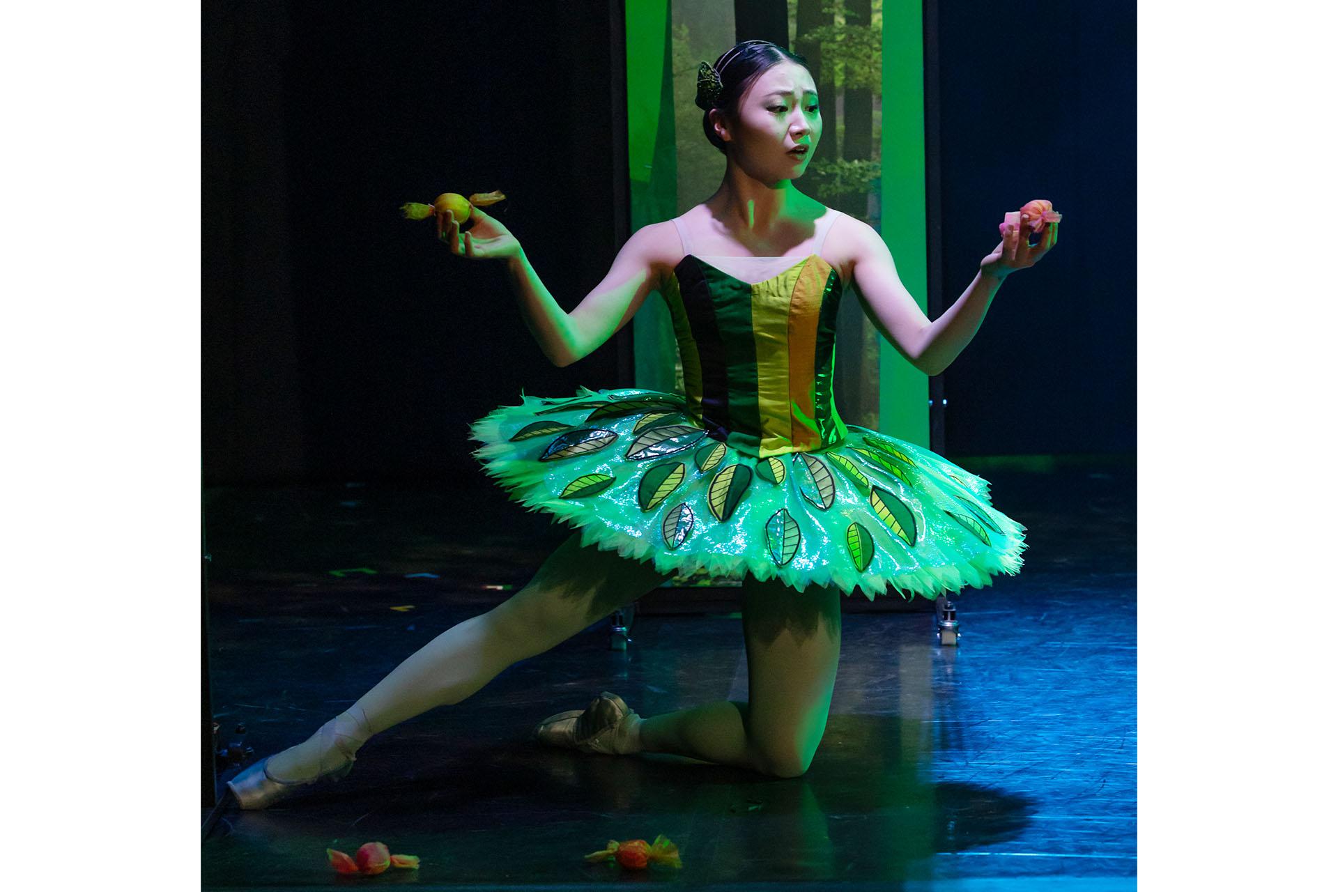 A female dancer in a green tutu with leaves attached is kneeling on the floor, picking up sweets. She looks shocked to see the sweets on the floor.
