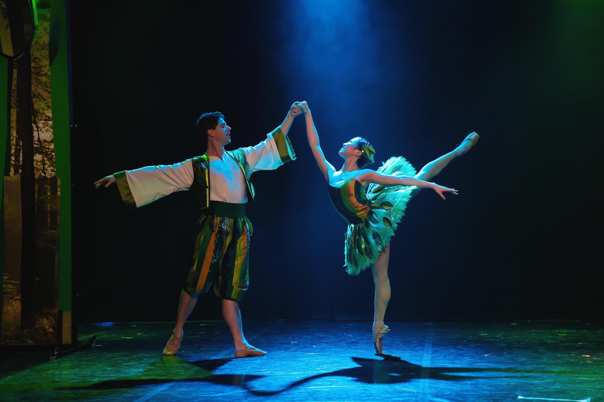 Two dancers stood in a traditional ballet pose. One dancer is on pointe with leg raised high