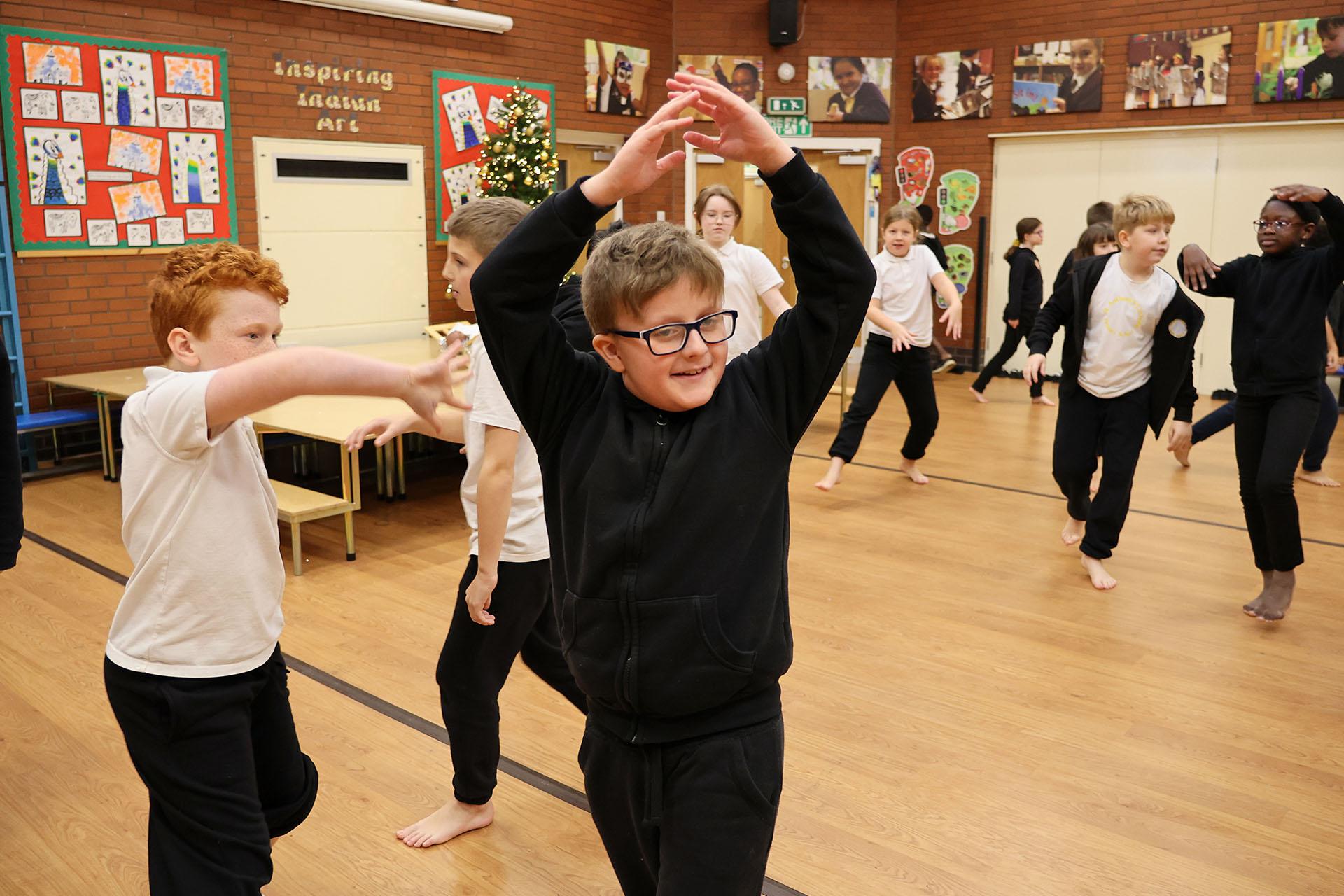 Schoolchild in a dark top and glasses holds their hands above their head, fingers touching, as he moves around the space
