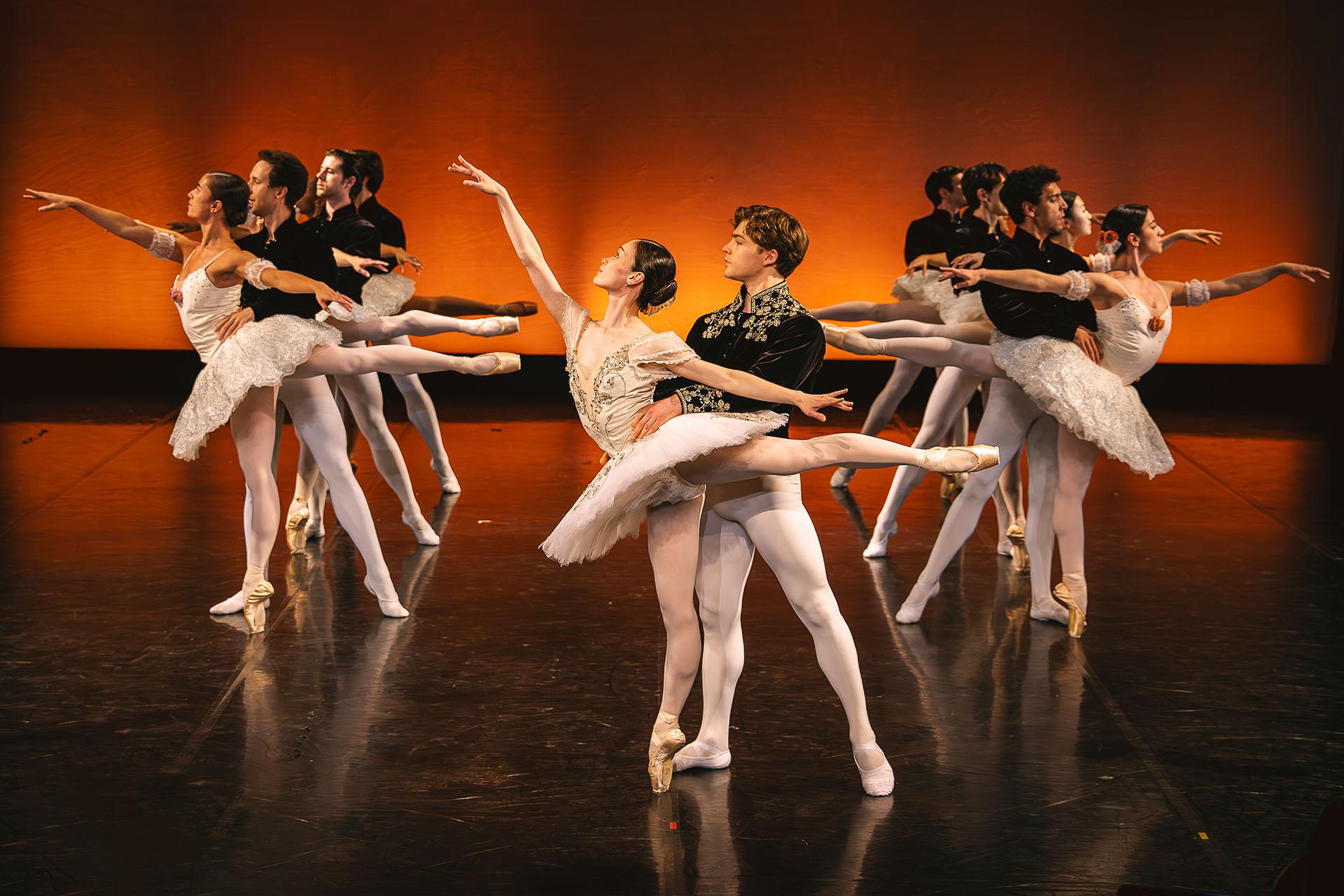 Seven pairs of dancers hold a pase where the man in a black top supports holding the waist of a female dancer en pointe standing arabesque