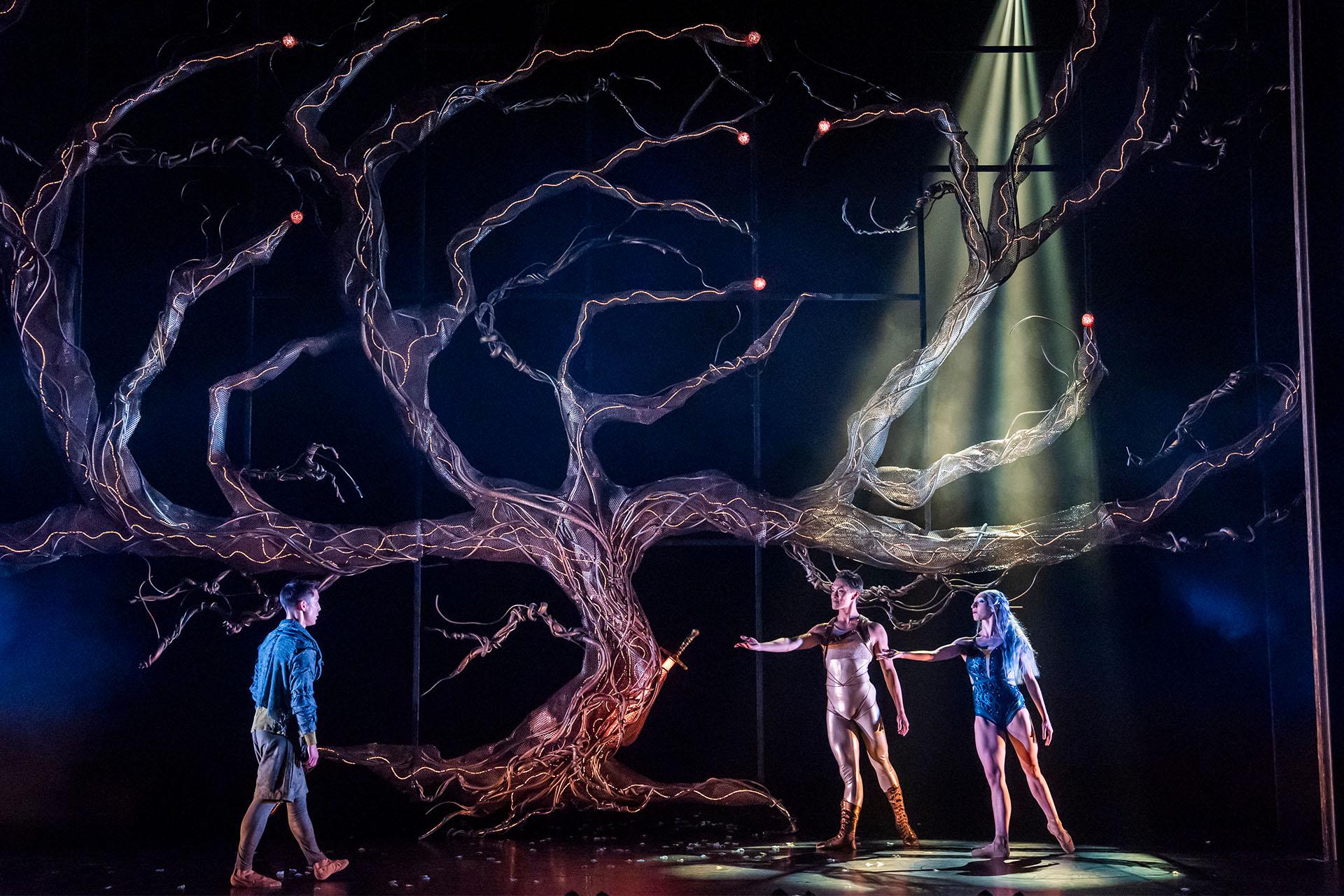 Under a leafless tree, Merlin approaches the beckoning Lady of the Lake wearing blue and Helios in golden reflective clothes.