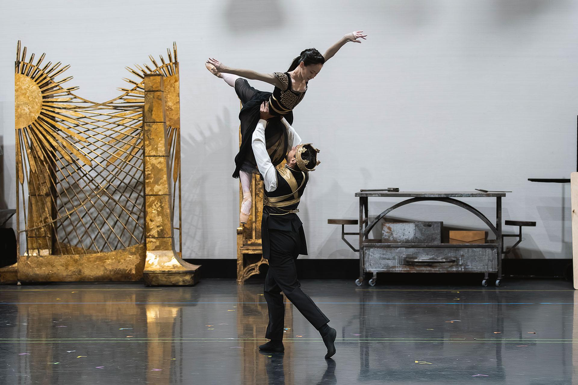 A man wearing a cron lifts a woman by the waist over in head, she has arms out, leg straight behind her and the other leg pointed straight down.