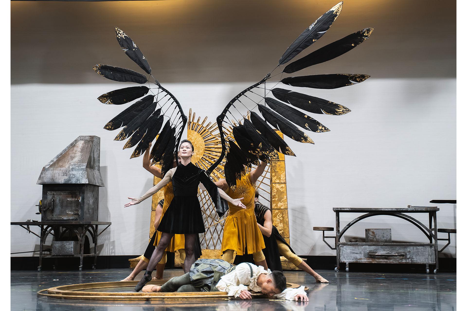 A woman in black with large black wings stands triumphantly over the prone body of a foe