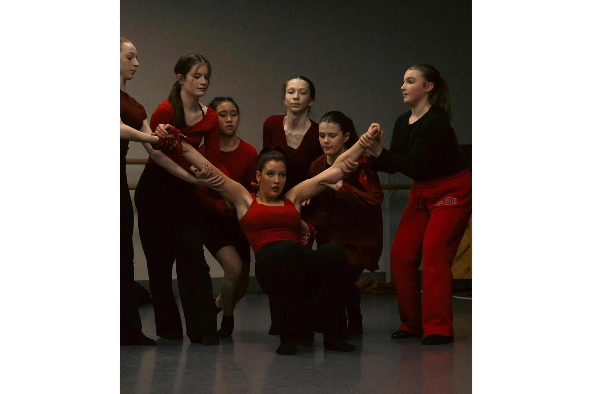Troop of dancers, one in a red top fallen backwards with those around her, also with red tops, holding her upper body off the dance floor