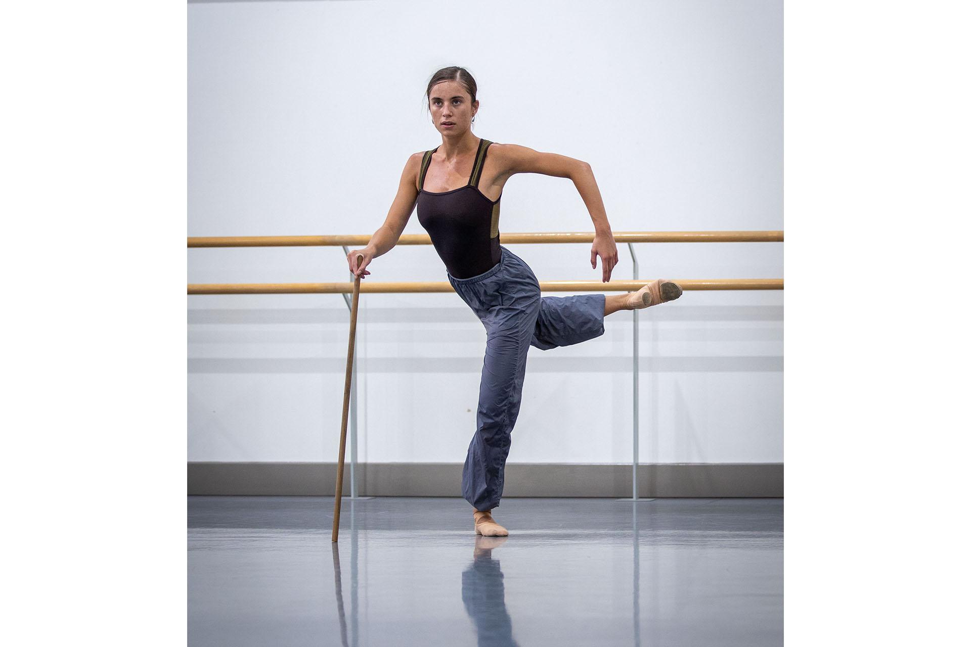 Dancer stands on one foot, a leg raised arabesque behind her, leaning forward on a walking stick