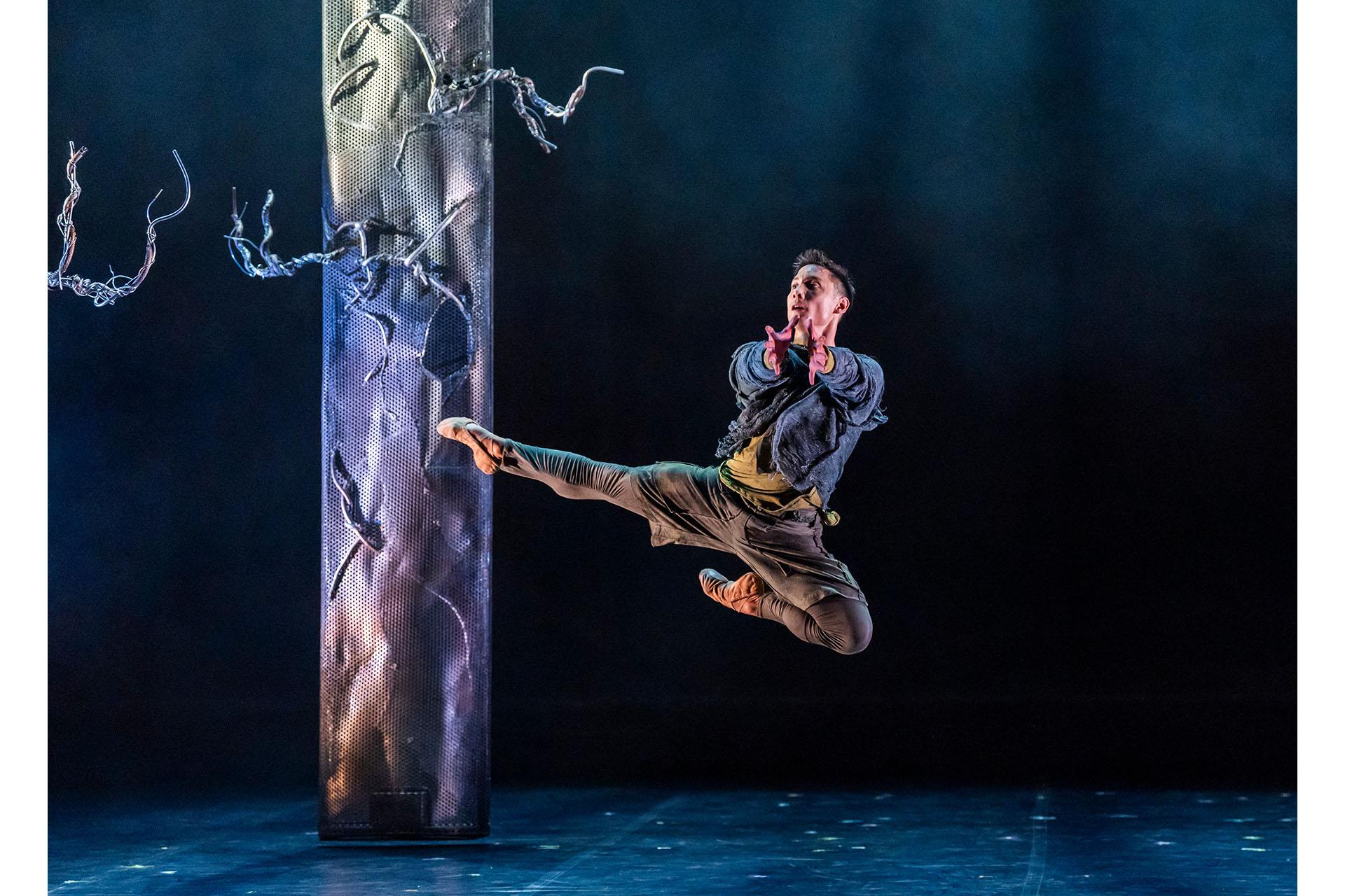 Merlin leaps in front of a tree, he is wearing simple, green, threadbare clothing