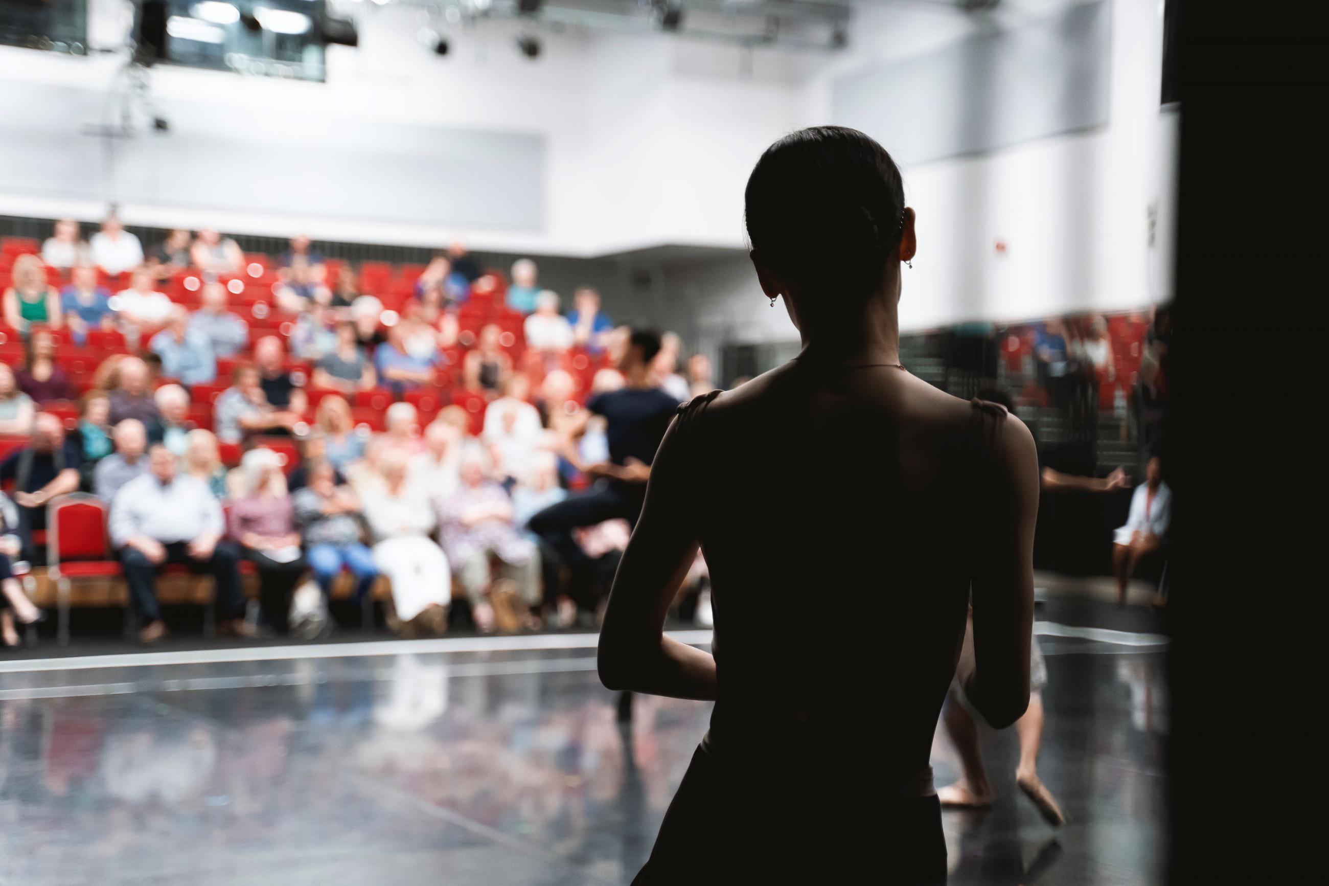 A silhouette of a dancer stood with their back to the camera while taking ballet class. On the background, a group of spectators watch from red chairs.