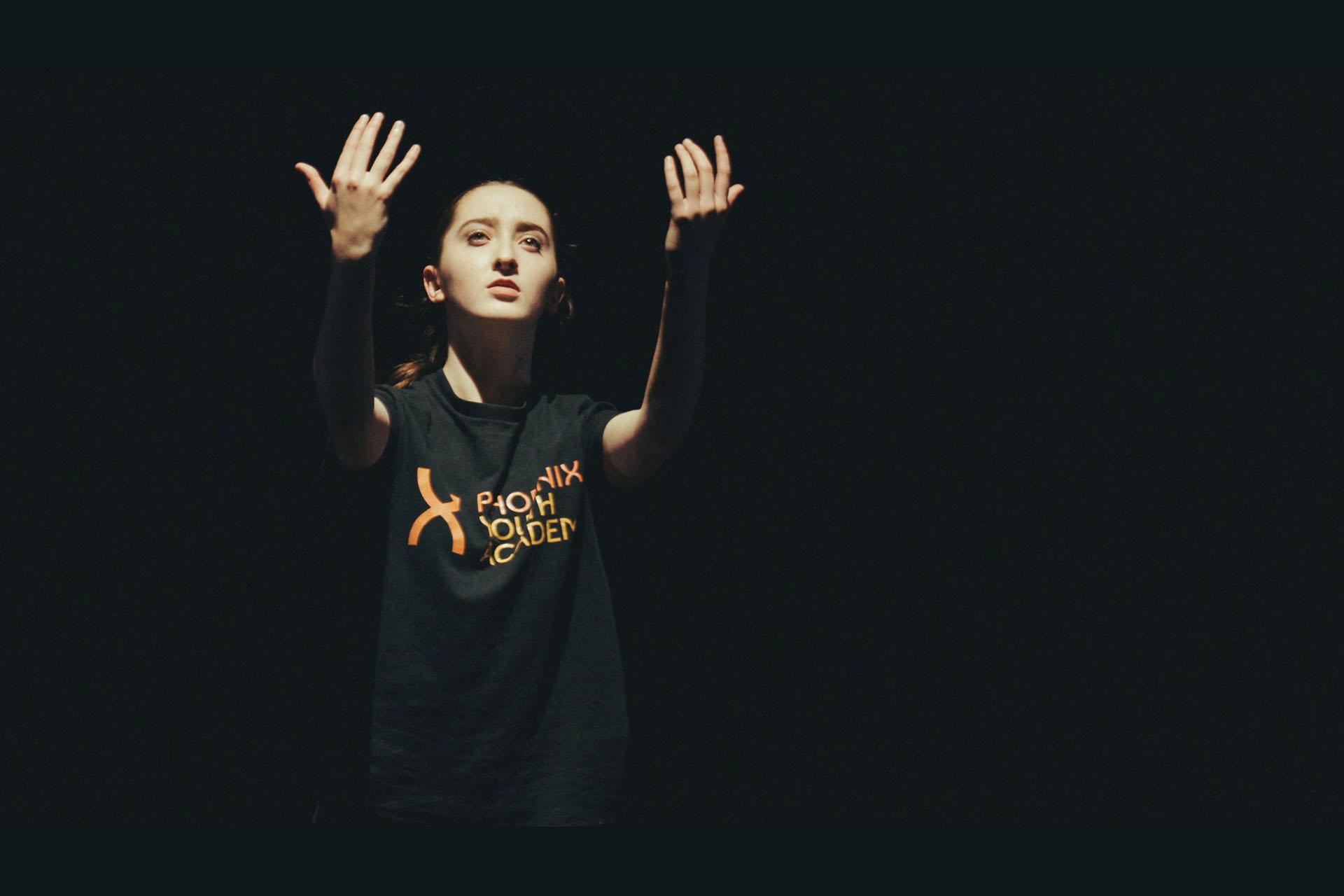 Student wearing a Phoenix Youth Academy t-shirt stands in the dark, single light illuminating them, arms forward and palms up