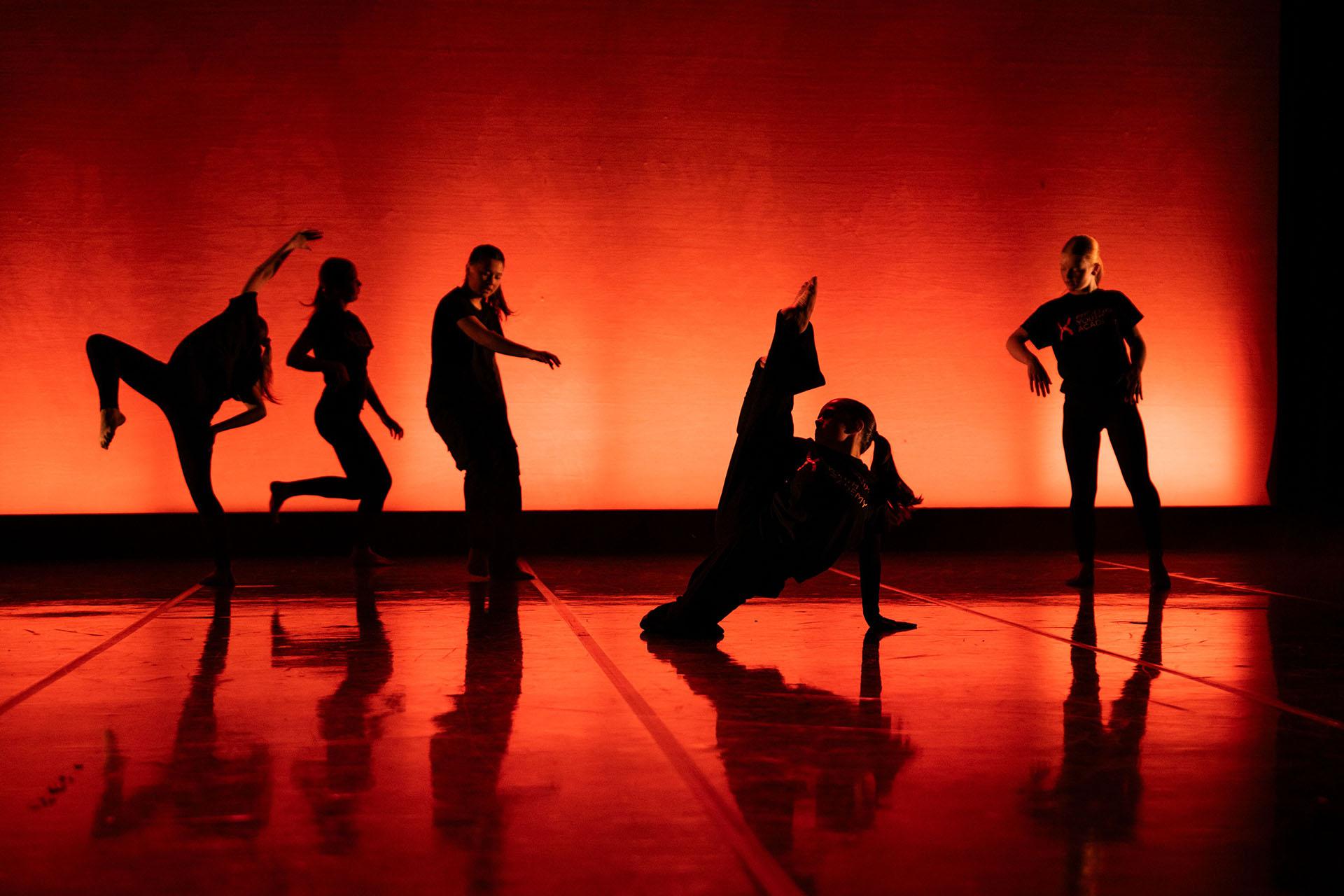 The back is illuminated in red and orange, dancers are closer to us in near silhouette, each in a different but striking pose